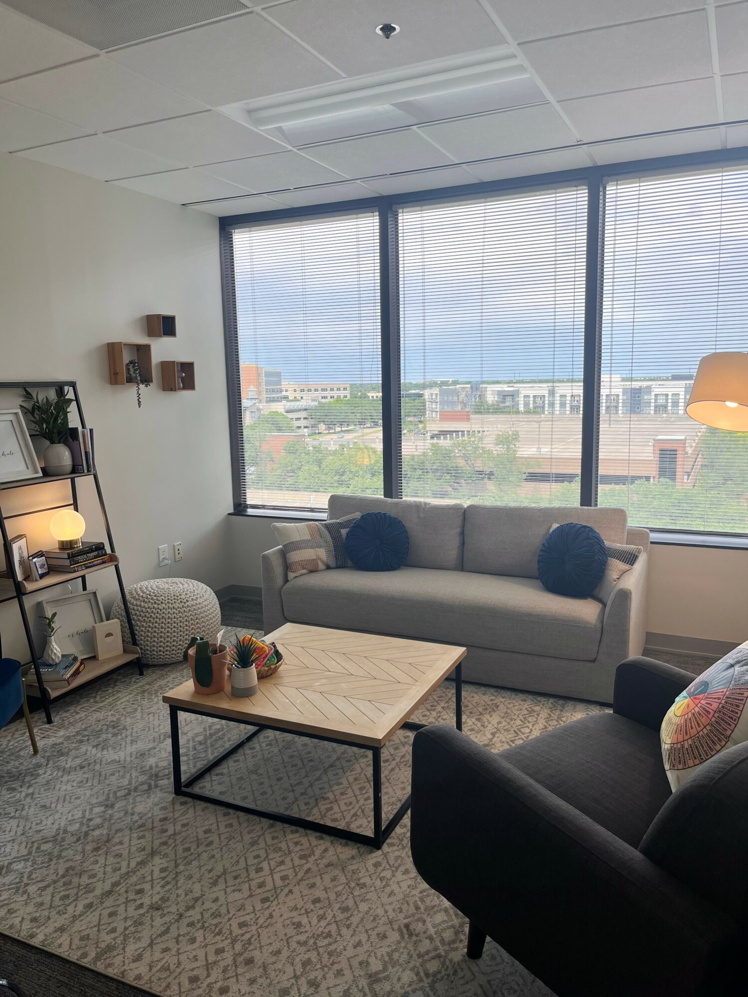 Living room with large window, sofa, armchair, coffee table, and bookshelf, overlooking cityscape.