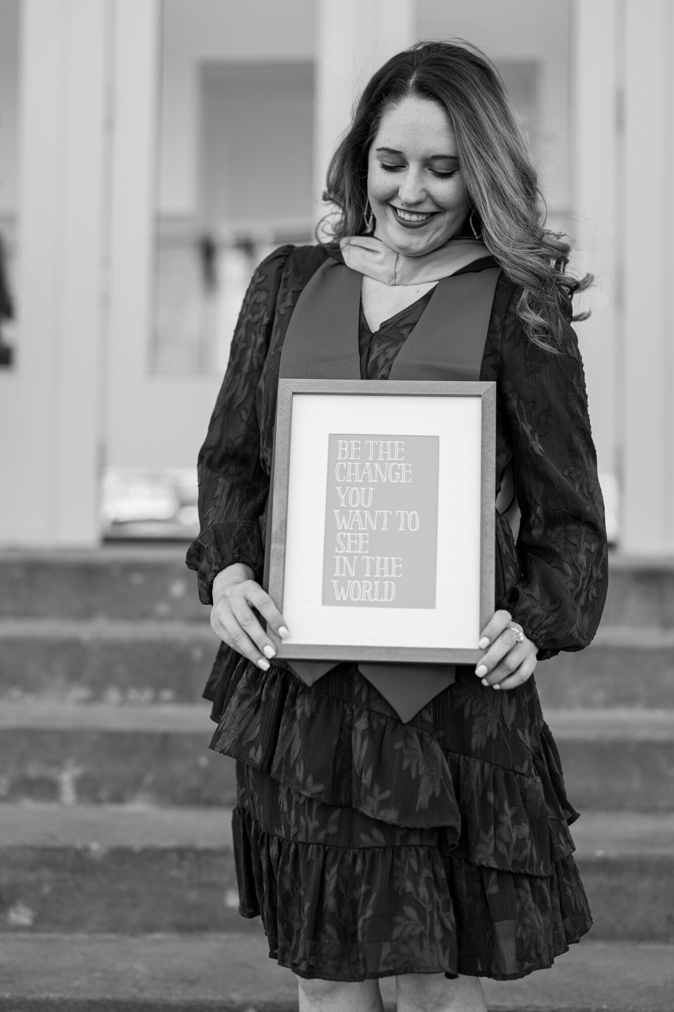 Woman in graduation gown holding a framed certificate, smiling outdoors on steps, in black and white.
