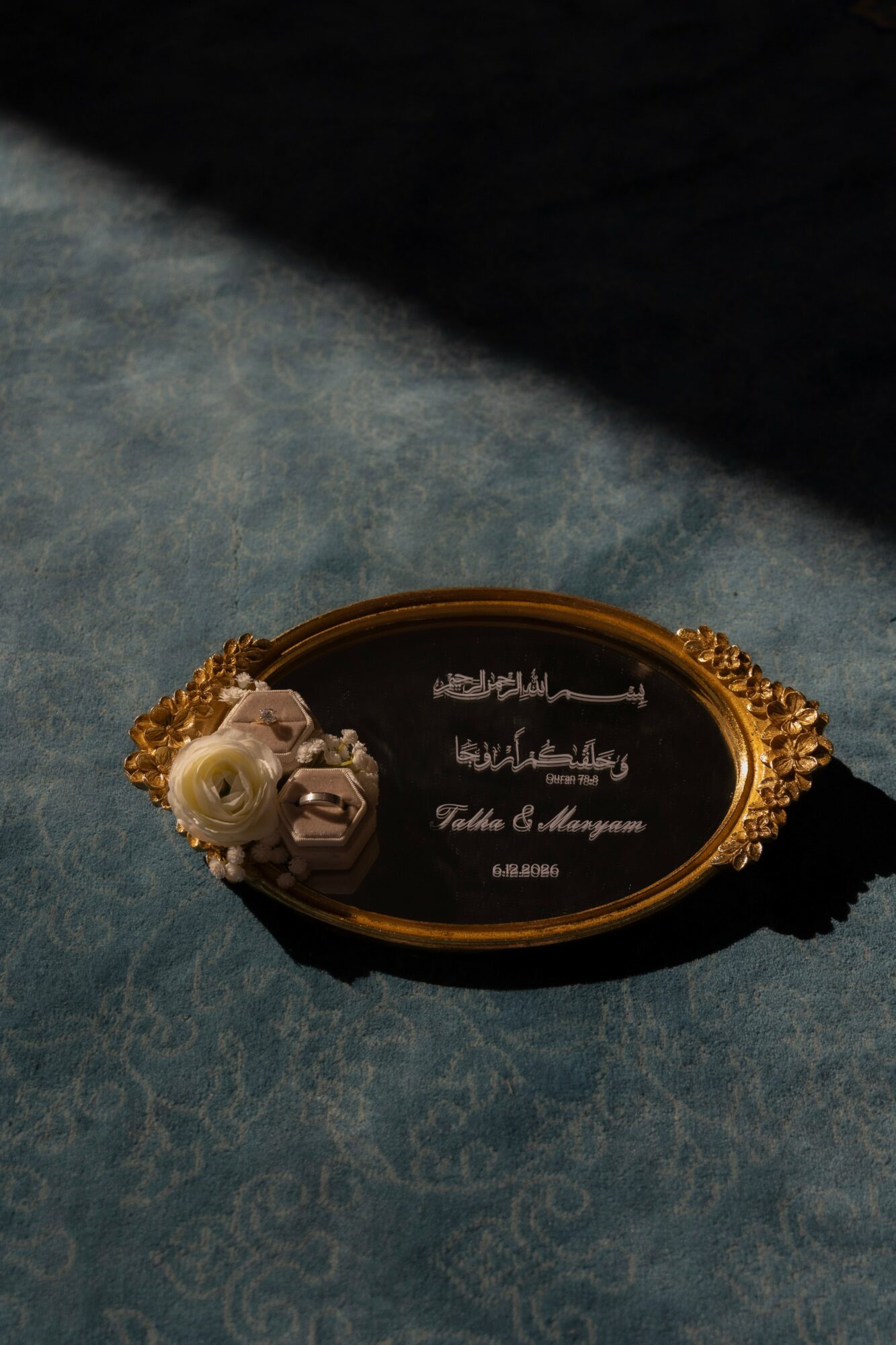 Decorative black and gold plaque with Arabic script and a white flower, placed on a textured surface.