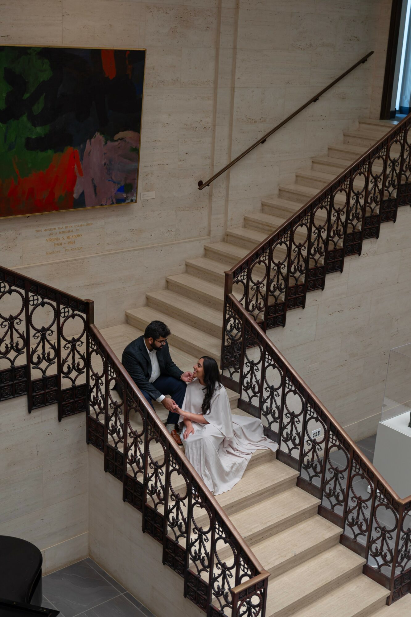 Two people sitting on a staircase railing in a building interior, one in a dark suit and the other in a white dress, with a painting on the wall behind them.