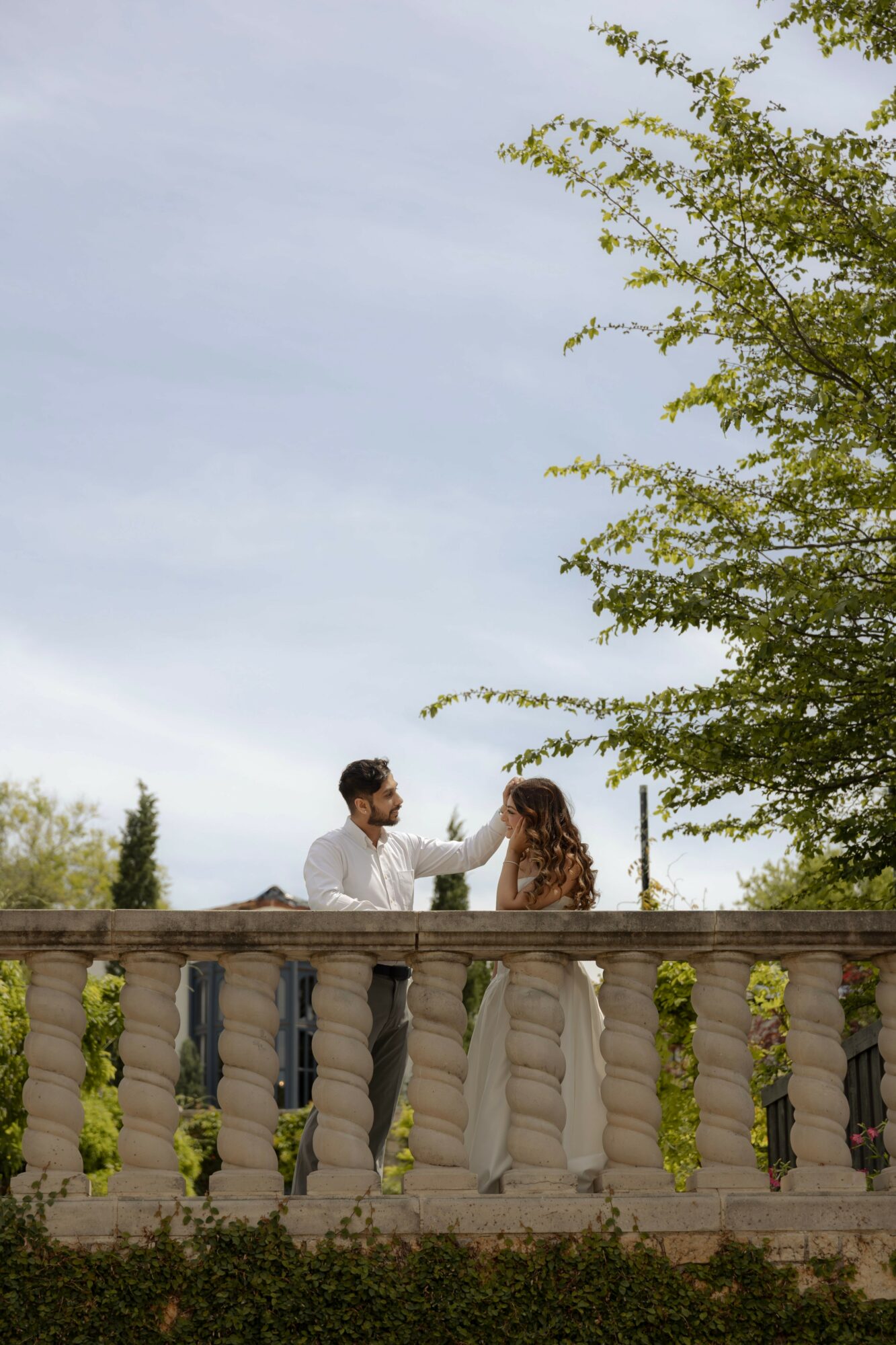 A man and woman stand on a balcony with a stone railing, surrounded by trees and a cloudy sky.