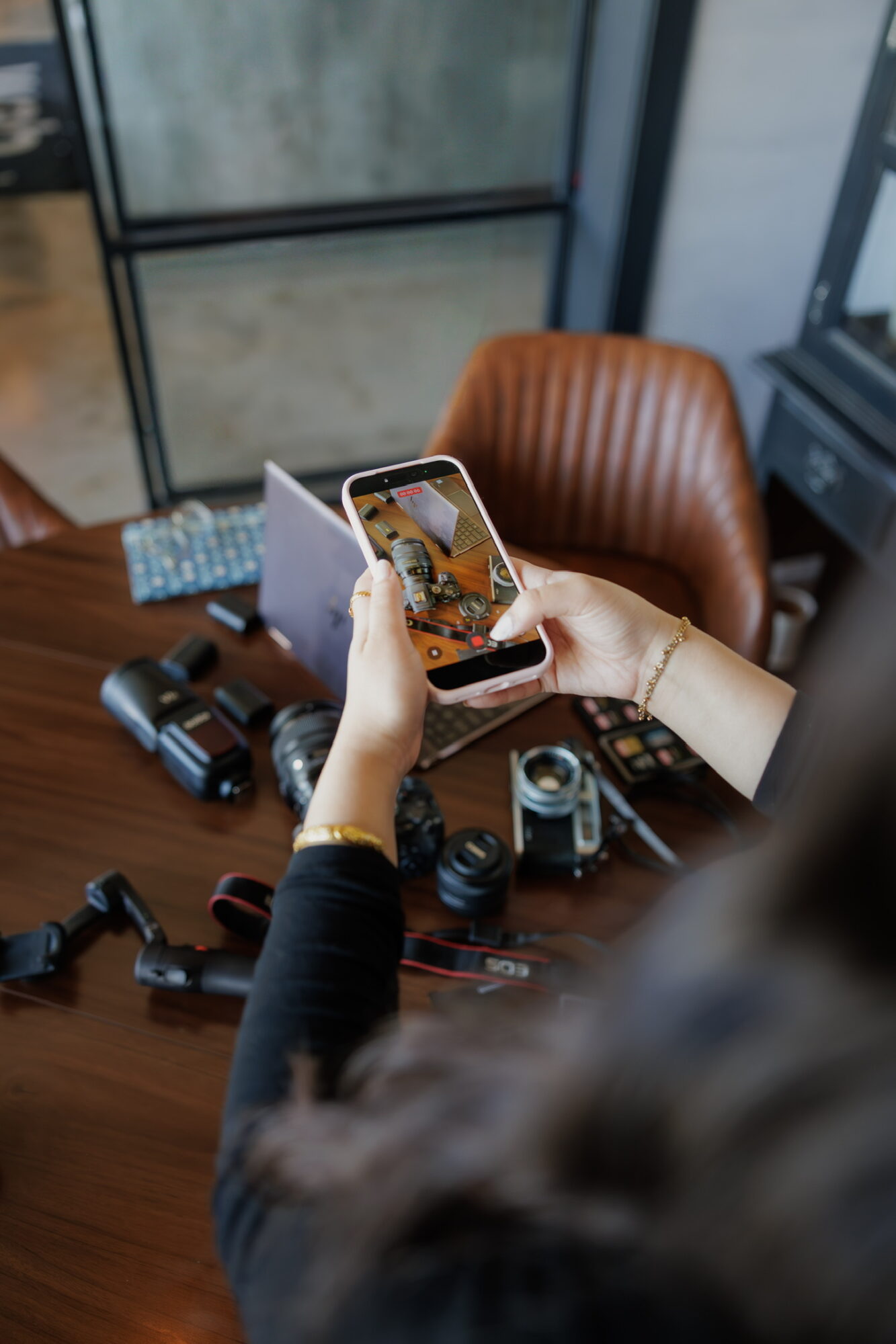 Person taking a photo with a smartphone of a camera on a table, in a room with large windows and a brown chair.