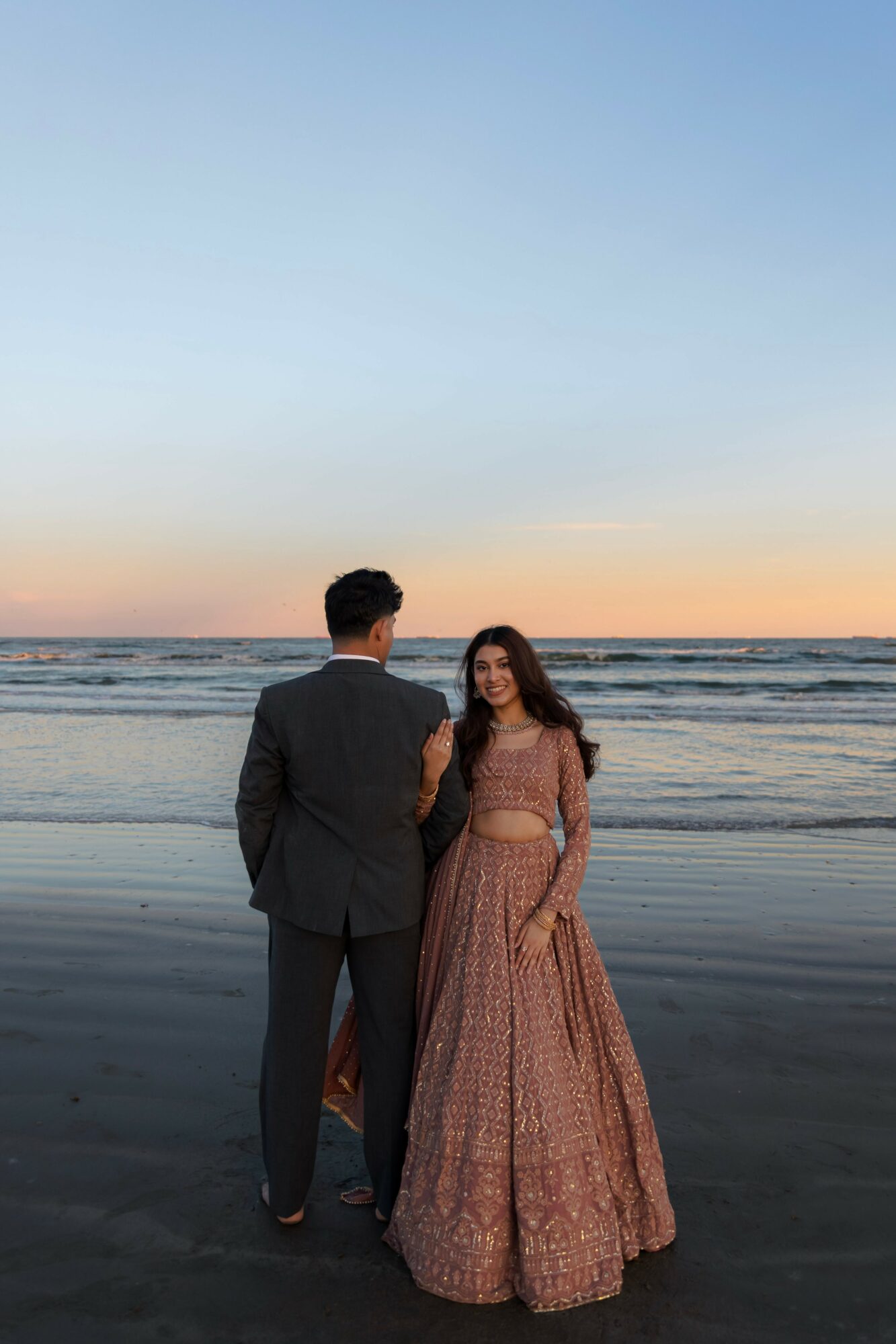A man and woman stand on a beach at sunset, facing the ocean, with the sky transitioning from blue to orange.