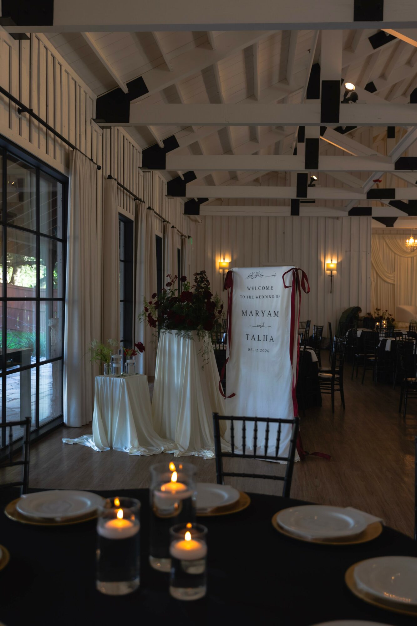 Indoor event space with decorated tables, chairs, and a floral arrangement, illuminated by warm lighting, with a high ceiling and wooden floor.