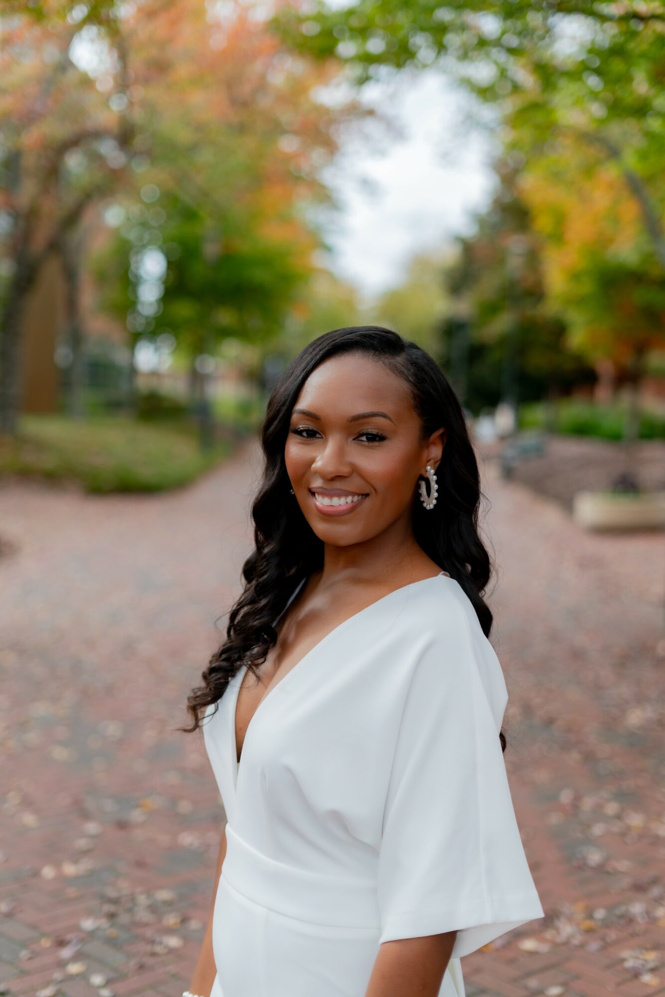 Smiling woman with long dark hair in a white dress outdoors during autumn, trees with colorful leaves in background.