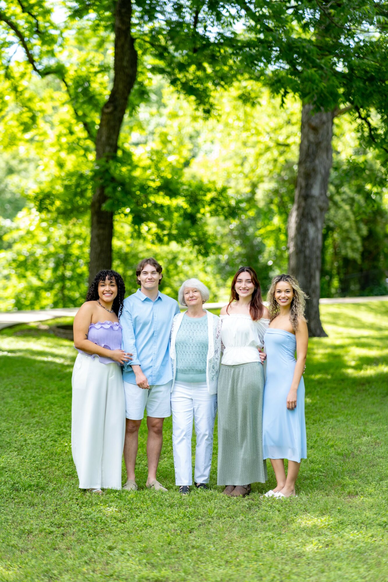 Group of five people standing outdoors on grass with trees in background, smiling, dressed in casual summer clothing.
