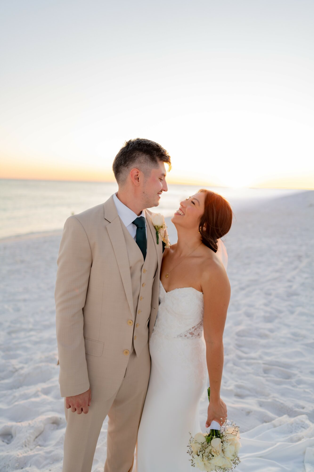 A bride and groom stand on a beach at sunset, looking at each other, with the ocean and sky in the background.