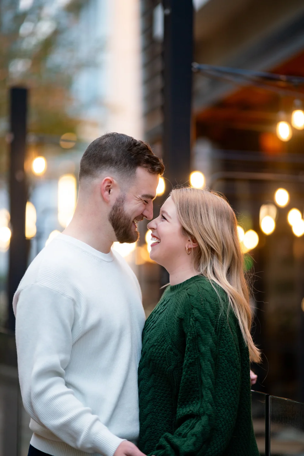 A smiling couple faces each other outdoors, with blurred lights and buildings in the background.