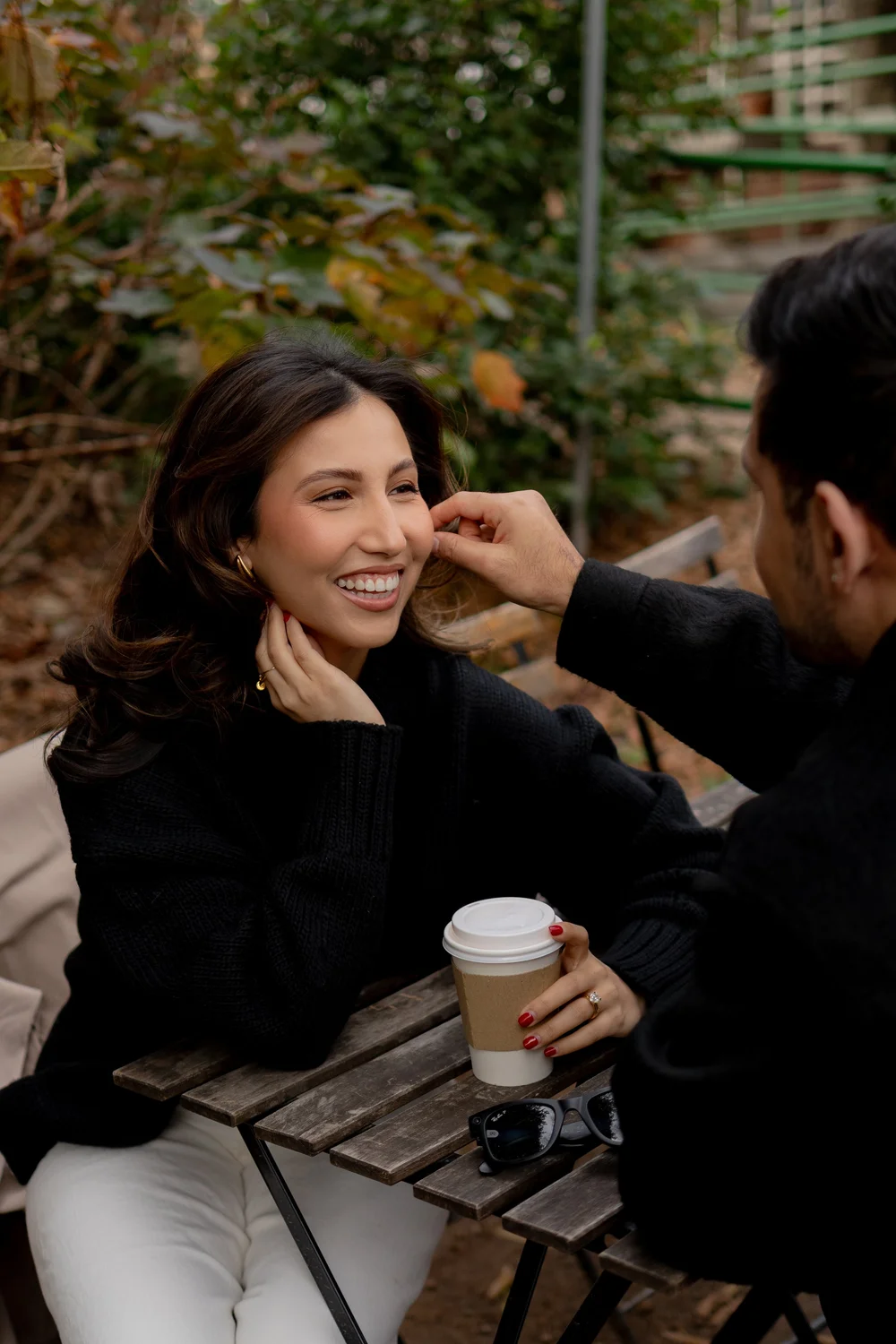 Woman smiling, holding a coffee cup, sitting at an outdoor table with a man touching her face, surrounded by greenery.