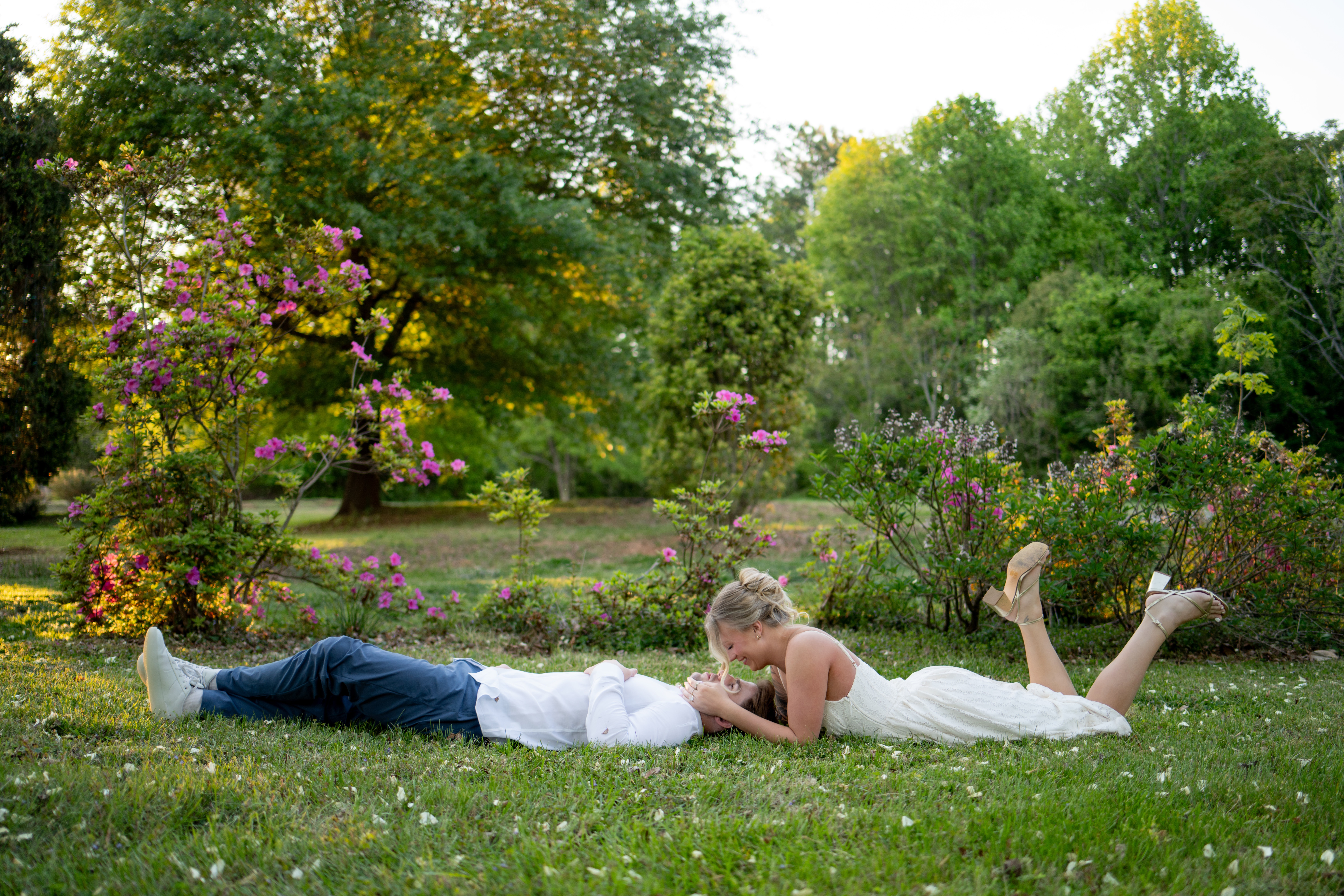 A woman and a man lie on grass in a park, facing each other, surrounded by trees and flowering bushes.