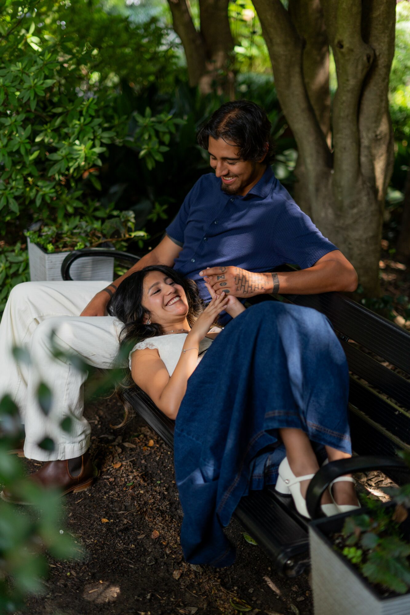 Two women outdoors, one sitting on a bench and the other lying on a park bench, smiling and interacting.