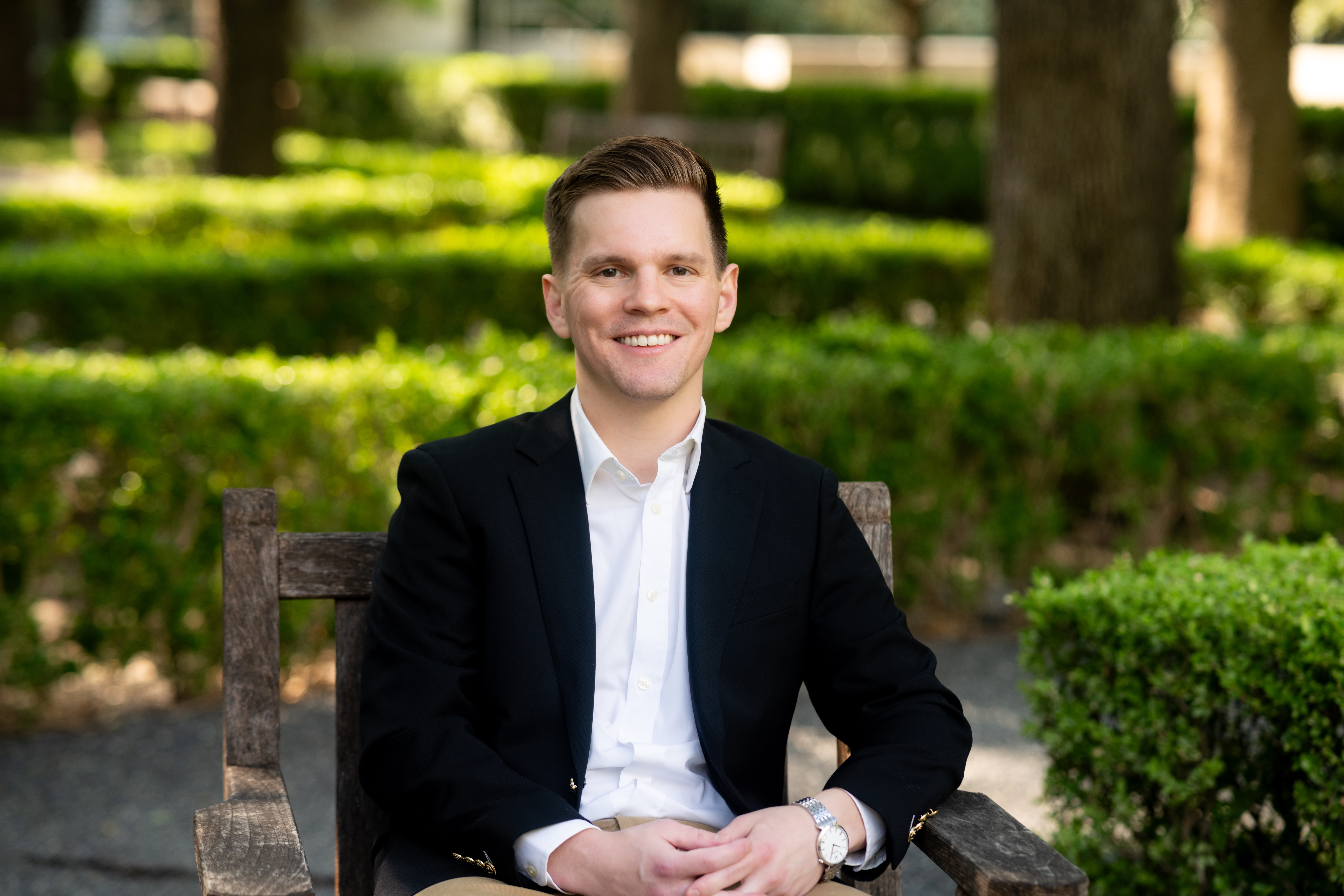 Young man in a suit sitting on a wooden bench outdoors, smiling, surrounded by greenery and trees.