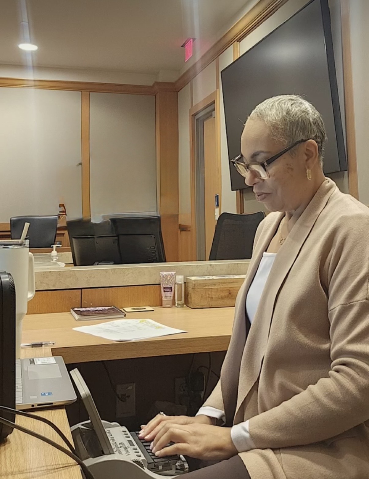 Woman with short hair and glasses working on a keyboard in a conference room with a large screen behind her.