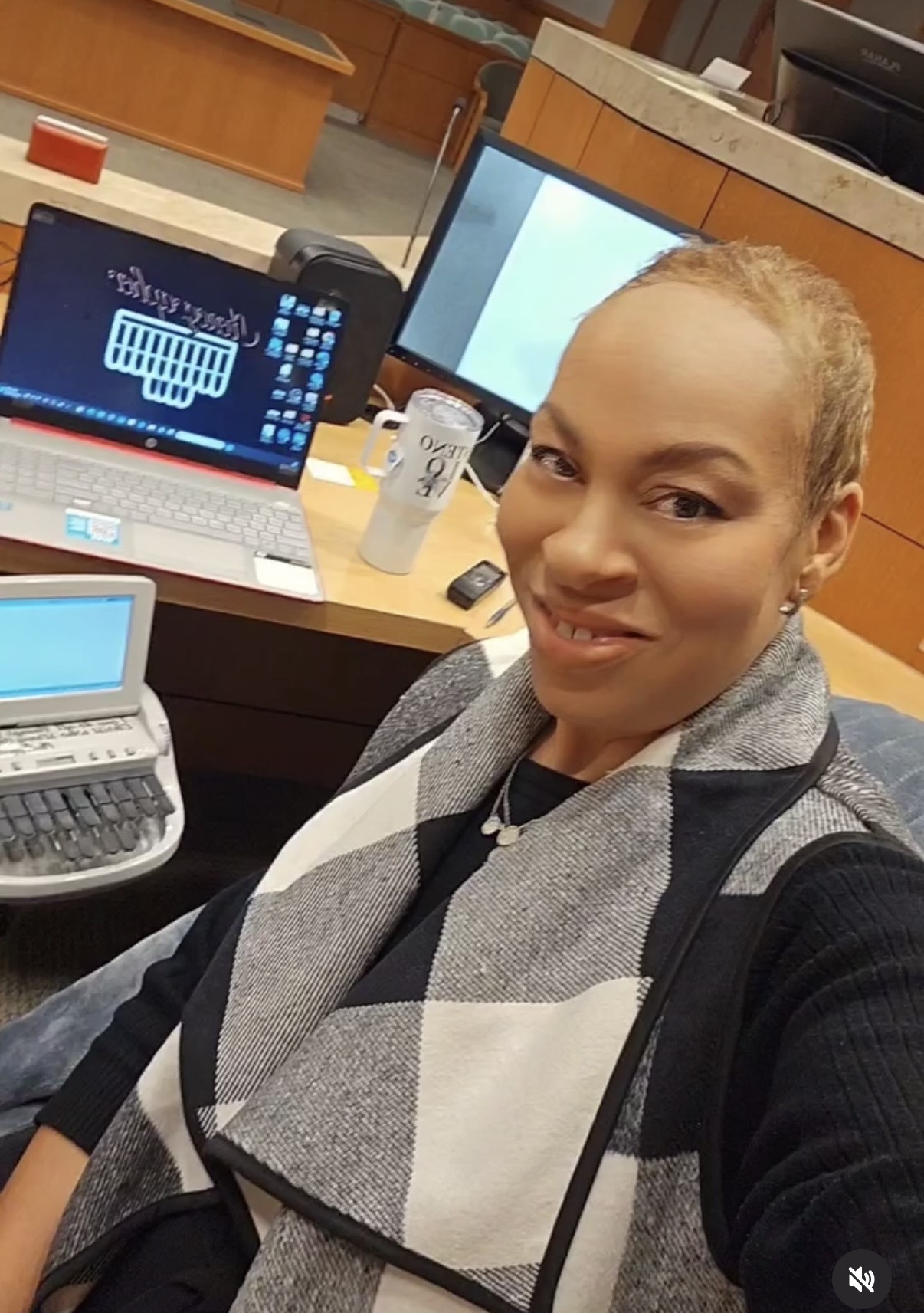 Woman with short hair taking a selfie in an office with computers and office supplies around her.