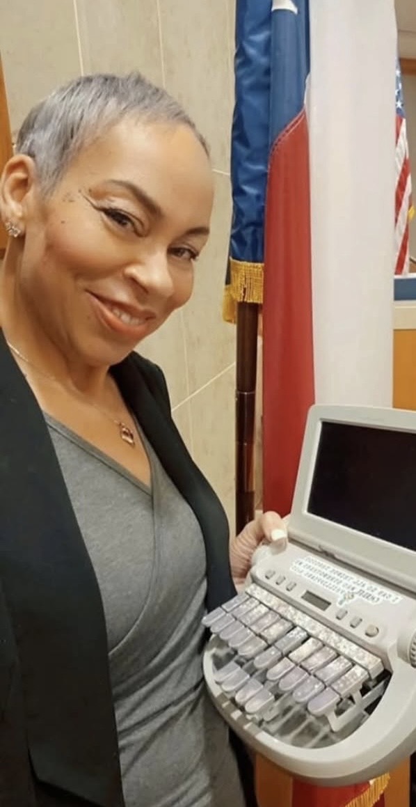 Woman with short gray hair smiling, holding medical equipment, in a room with flags and a computer screen.