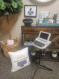 Office corner with a pillow, small monitor, basket, and plants on a wooden desk, against a wall with framed picture.