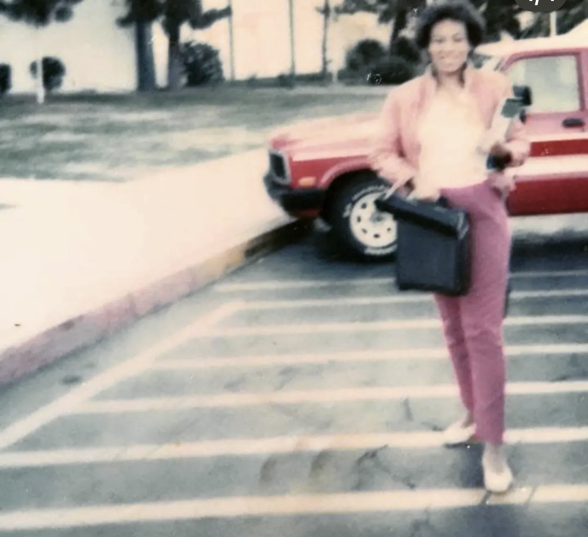 Woman walking in parking lot holding a black bag, red car in background, trees and buildings behind.