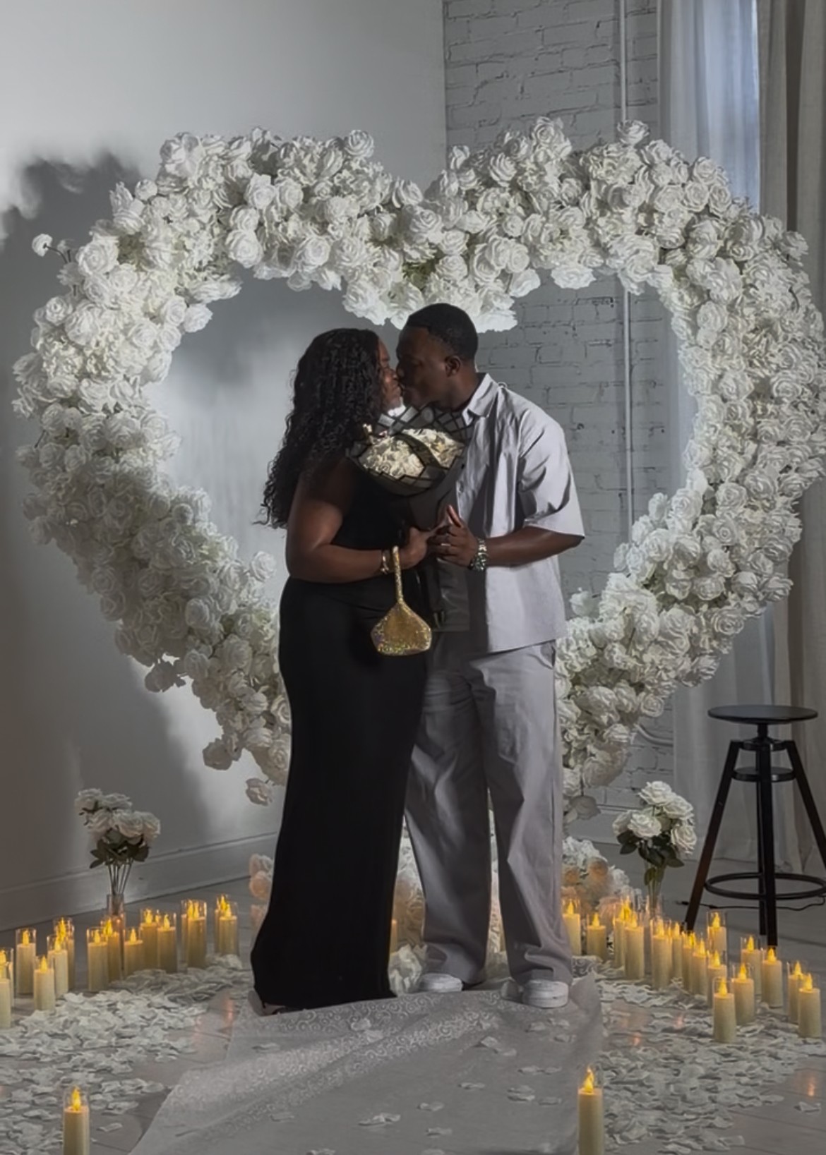 A couple stands inside a heart-shaped floral arch, holding a bouquet, with candles on the floor around them.