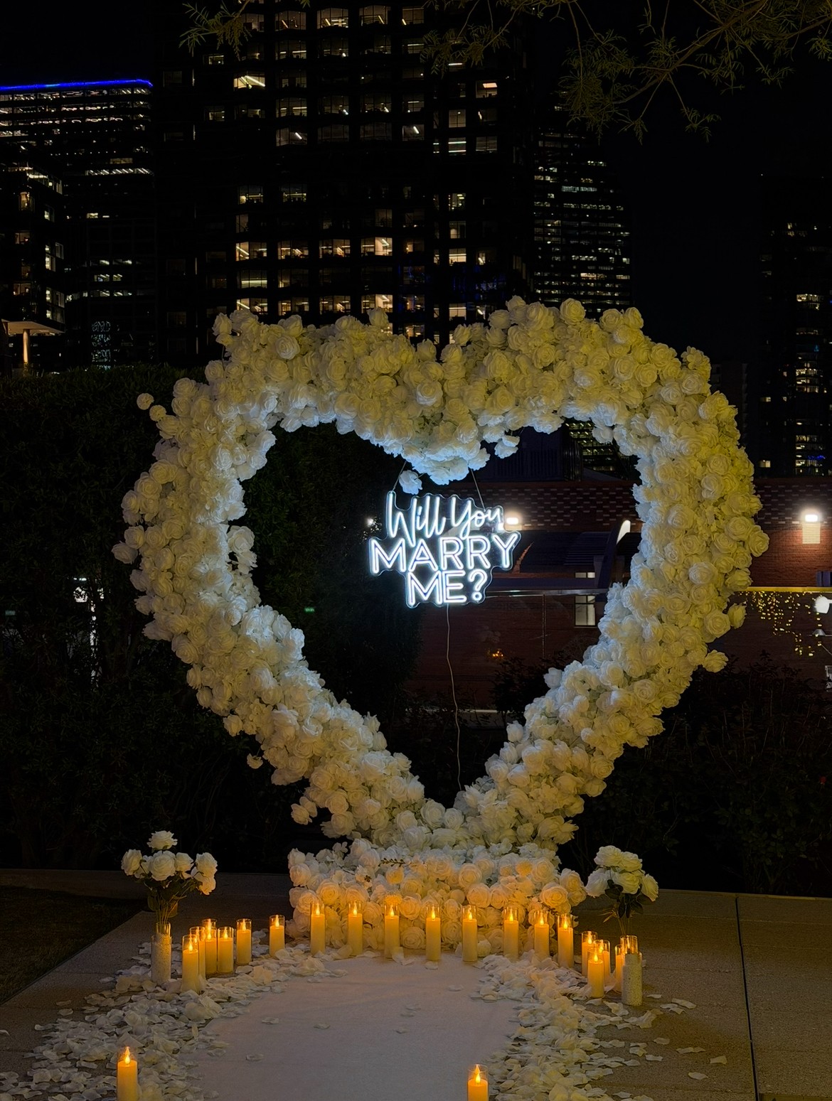 Heart-shaped floral arch with white flowers, candles, and a neon sign reading 'Will you marry me?' at night.
