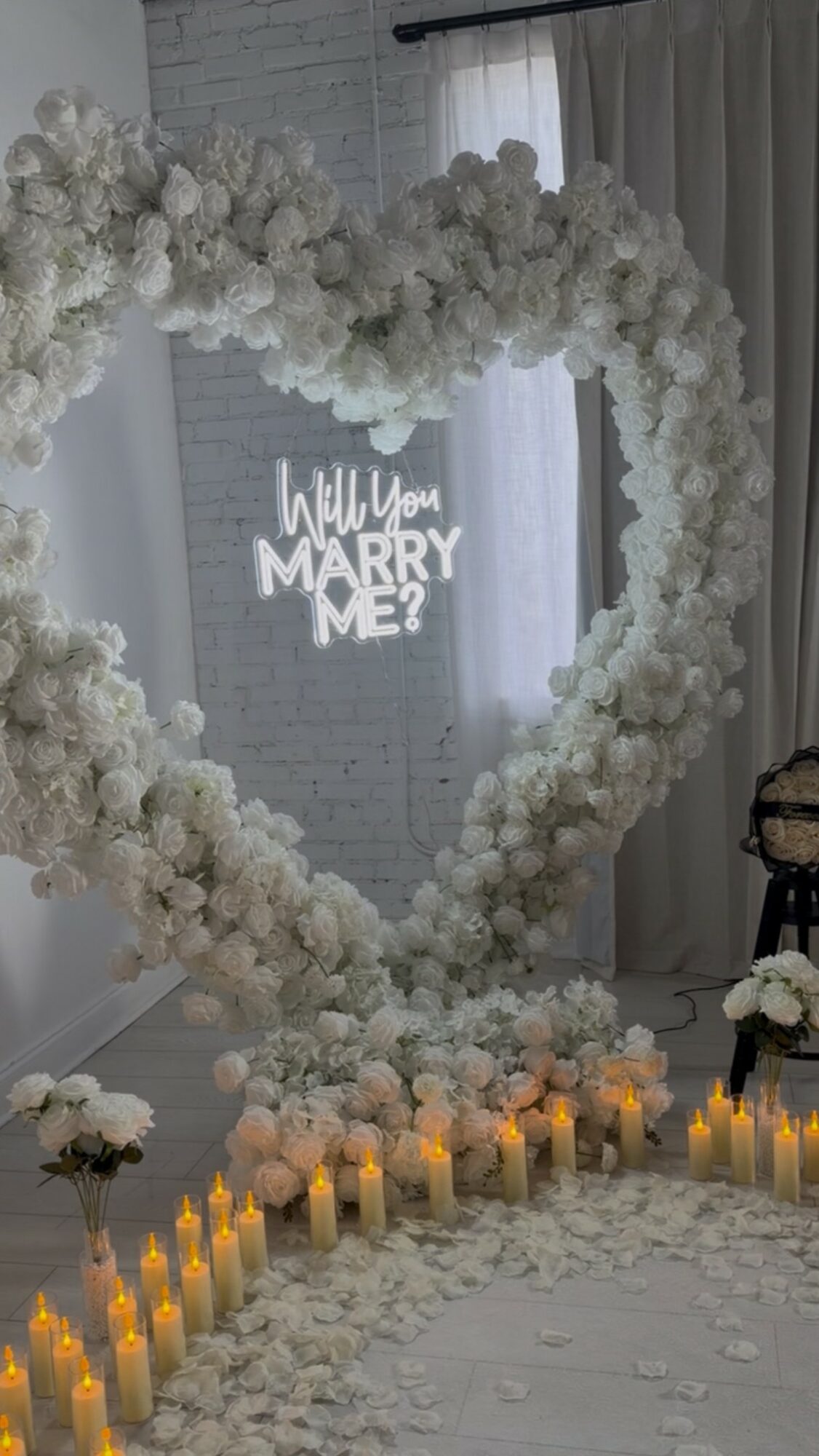 Heart-shaped floral arrangement with candles and a sign asking, 'Will you marry me?' in a decorated room.