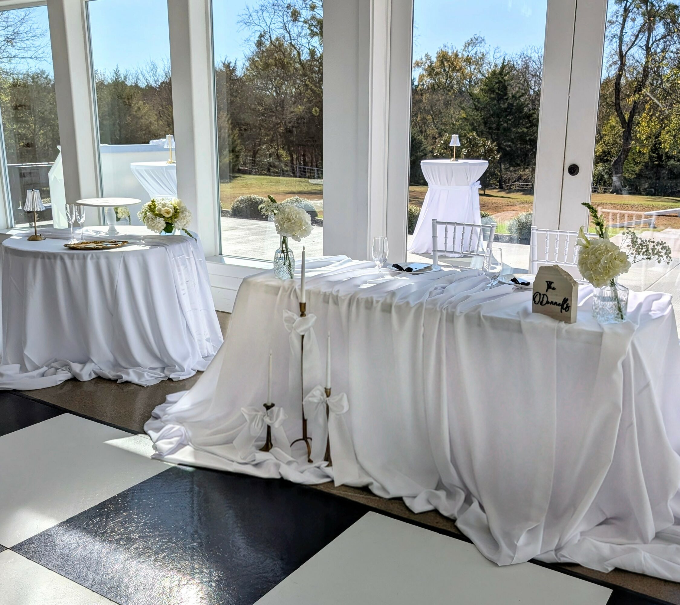 Table with white tablecloth and floral arrangements, set near large windows with outdoor view, sunlight streaming in.