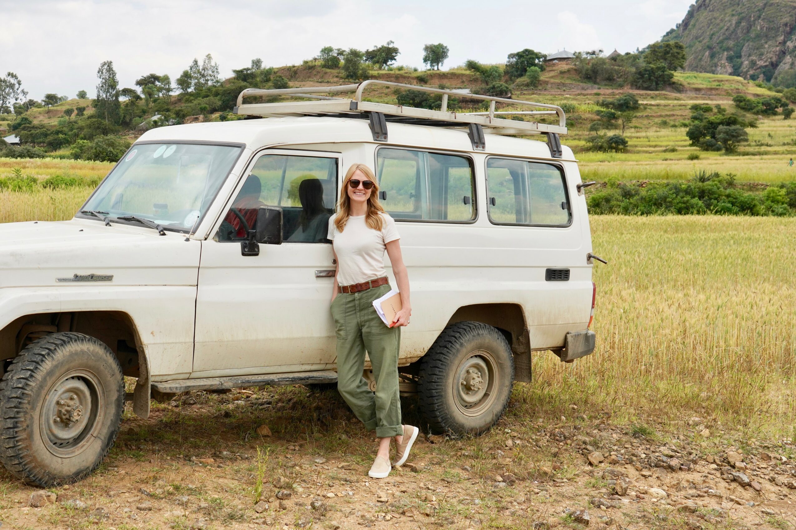 Woman with sunglasses standing next to a white off-road vehicle in a grassy field with hills in the background.