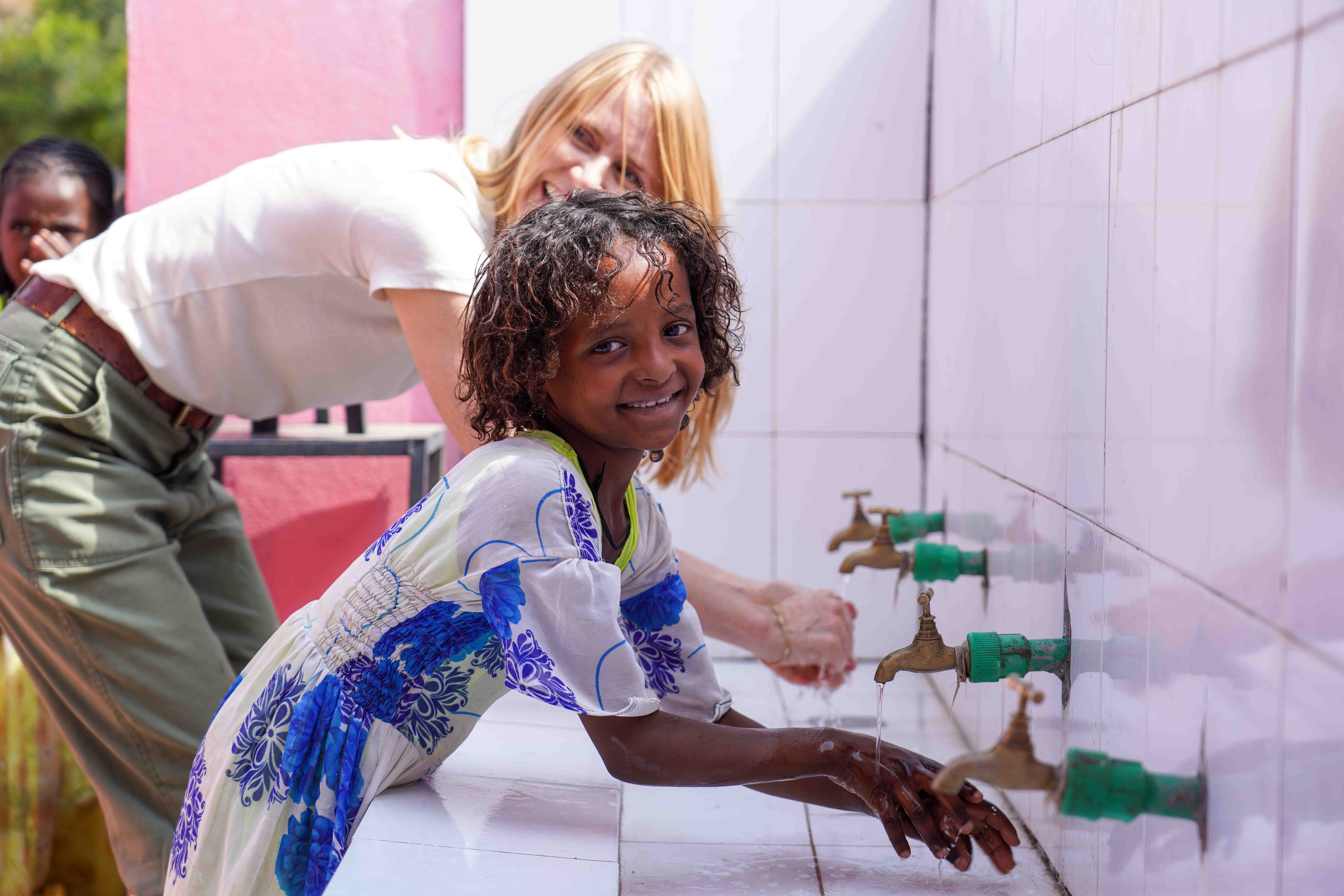 Child and adult washing hands at sinks with green faucets, smiling, in a tiled room.