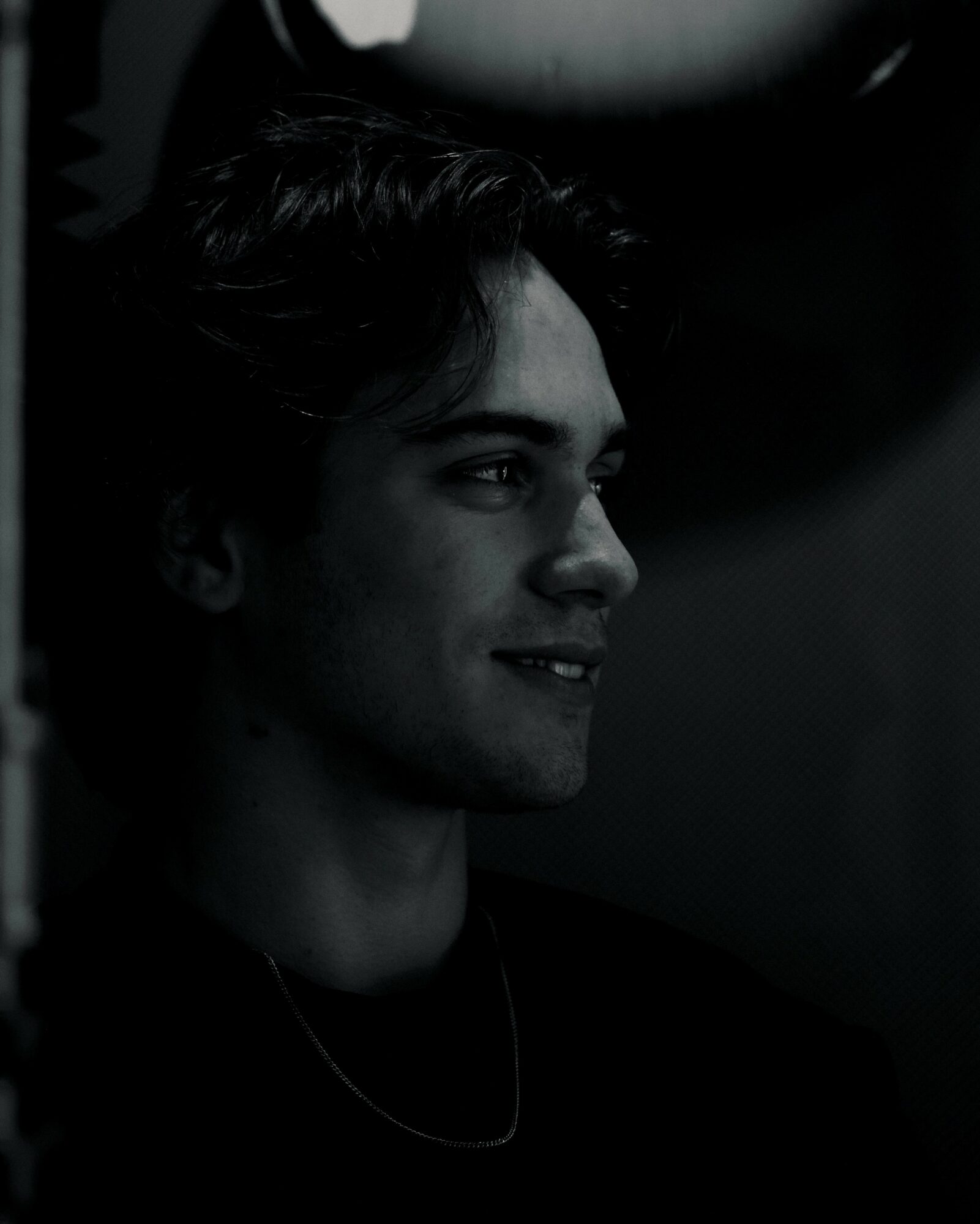 Young person with dark hair smiling, facing right, in a dark environment, with light highlighting their face.