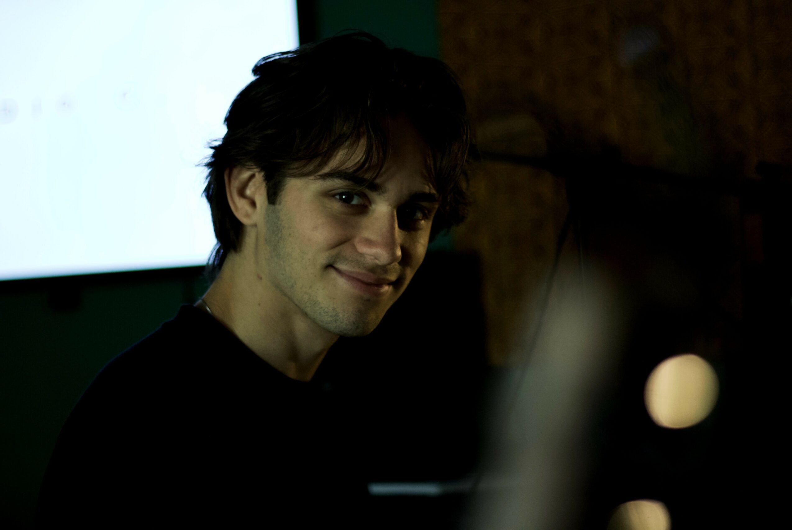 Young man with dark hair smiling in a dimly lit room, background includes a screen and wooden wall.