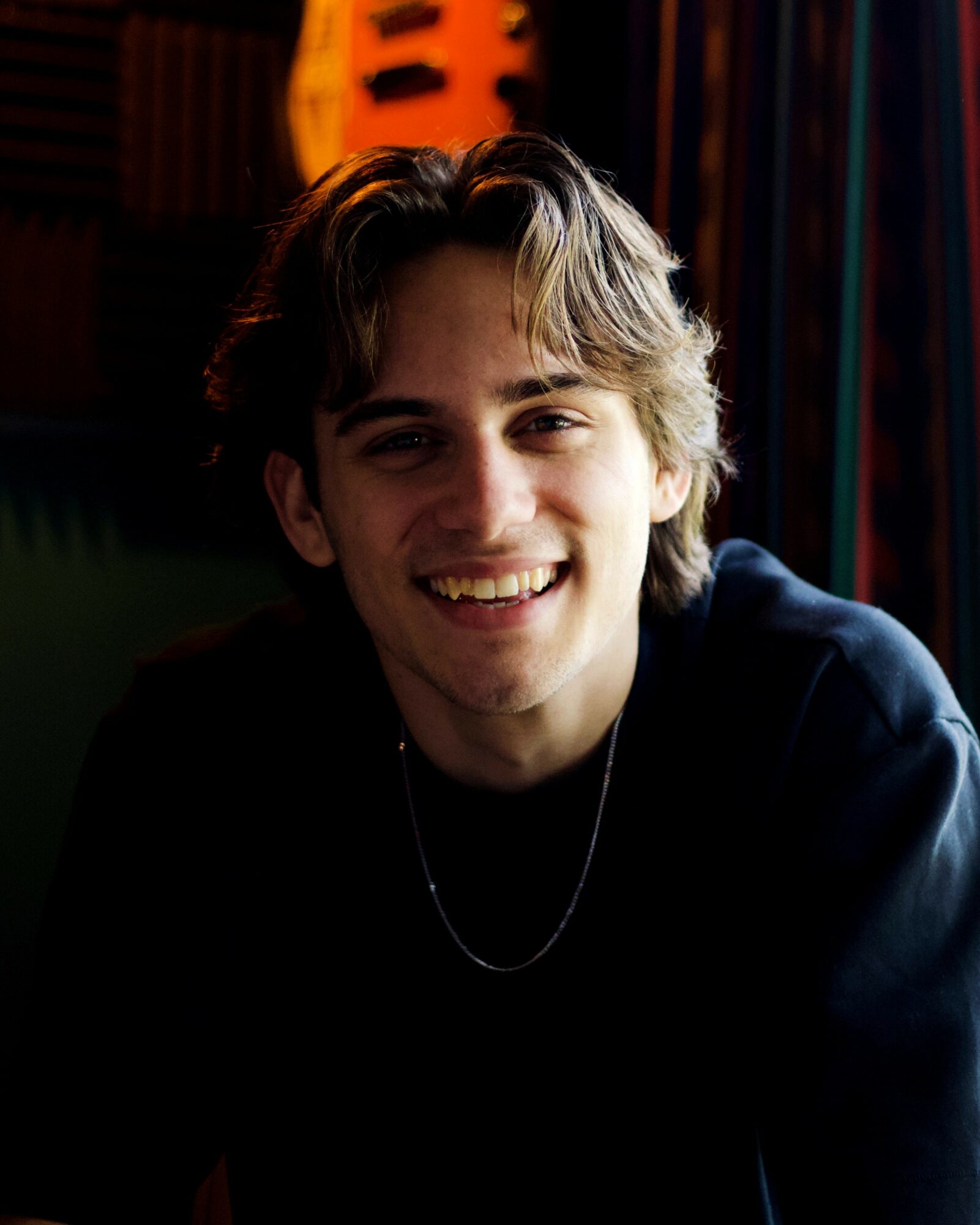 Young man with wavy hair smiling, wearing a dark shirt, in a dimly lit room with colorful background.