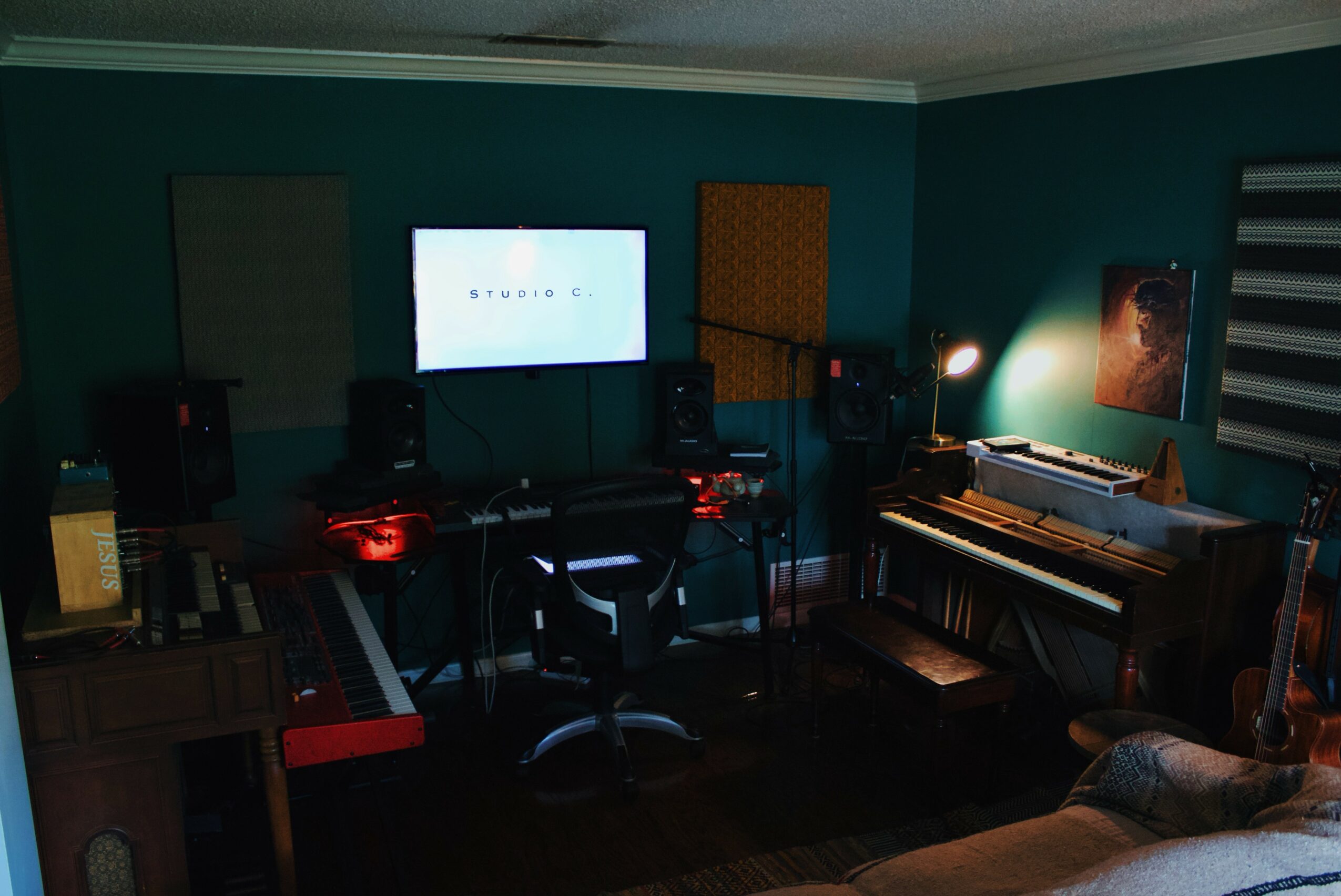 Music studio with computer, keyboard, speakers, and a wall-mounted monitor in a teal room.