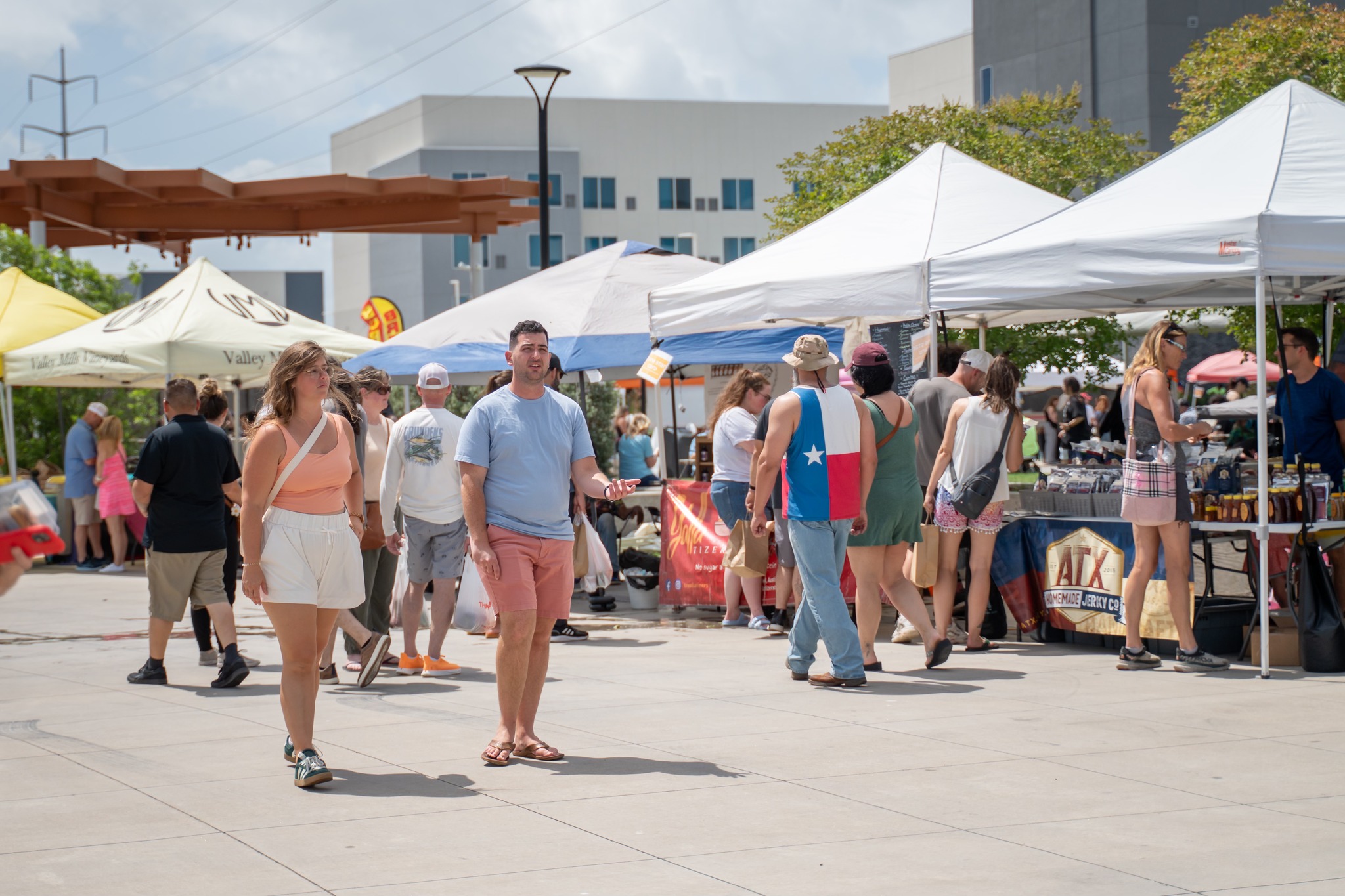 People browsing outdoor market stalls with tents, in an open urban area, sunny day, modern buildings in background.