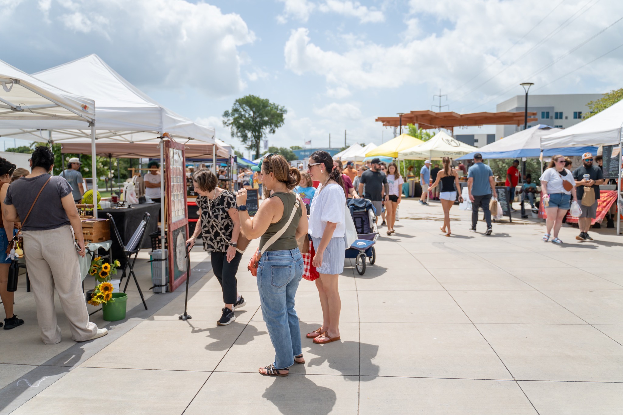 People browsing outdoor market stalls with tents and umbrellas on a sunny day, some walking and shopping.
