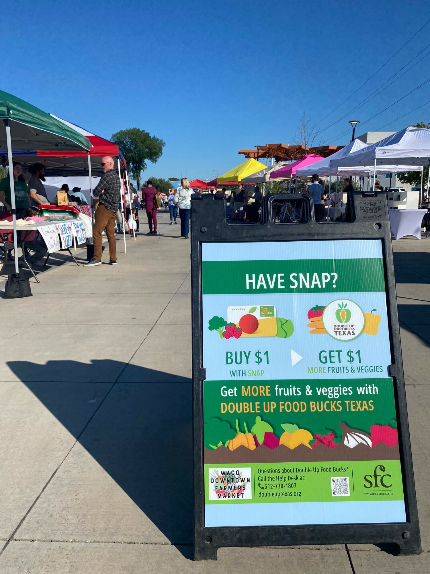 Outdoor market with a sign advertising a promotion for a mobile app, people walking, tents, and a clear blue sky.