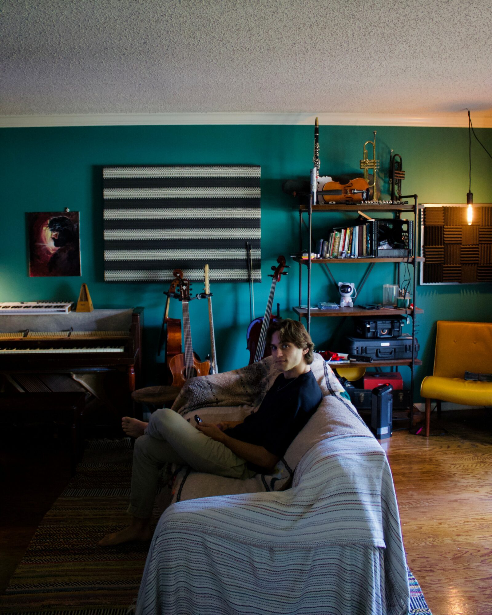 Person sitting on a sofa in a living room with teal walls, bookshelves, and a yellow chair, holding a phone.
