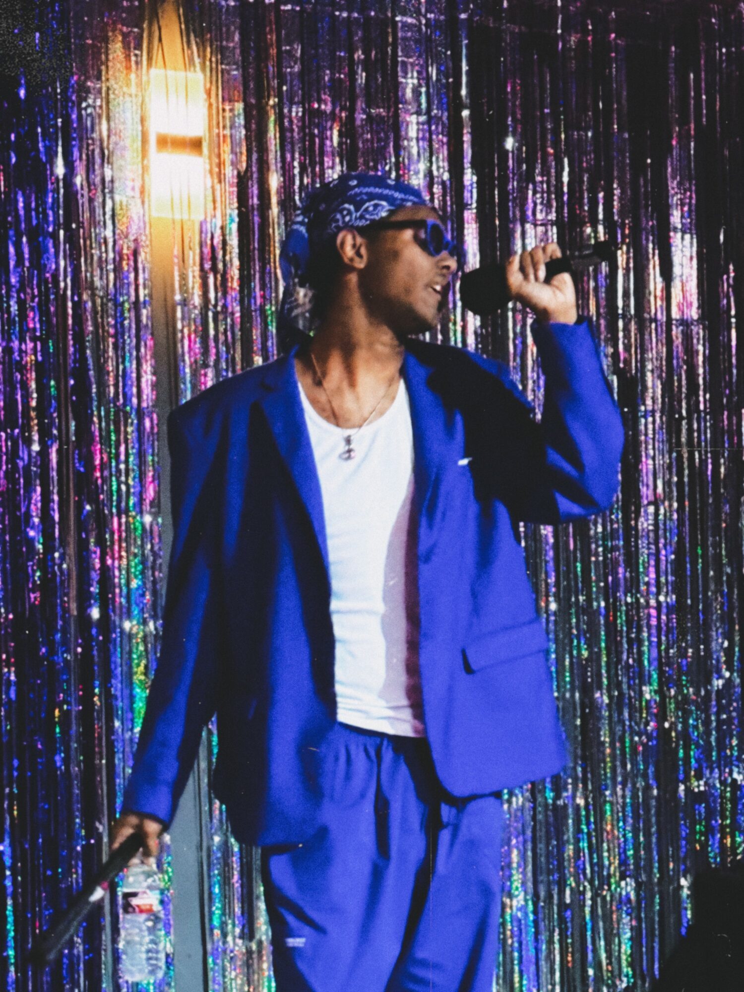 Person in blue suit and sunglasses singing into a microphone on a colorful, shiny backdrop.