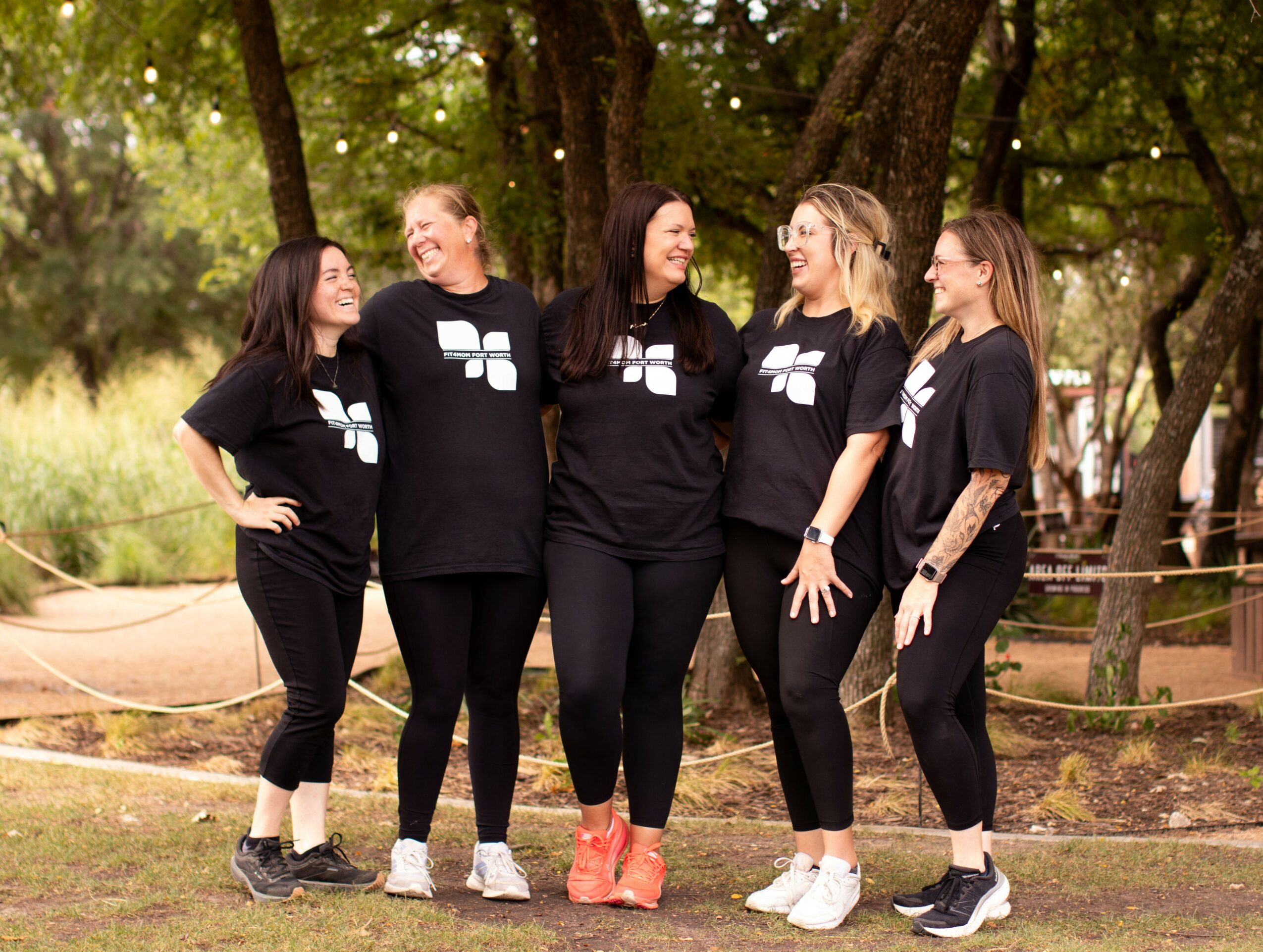 Five women stand outdoors, smiling and talking, surrounded by trees and grass, wearing black t-shirts with white logos.