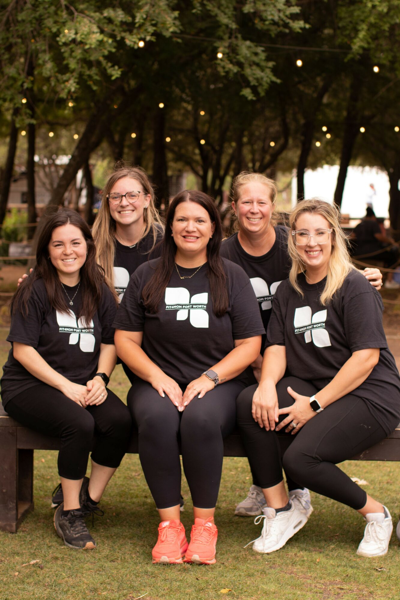 Five women sitting outdoors on a bench, smiling, with trees and string lights in the background.