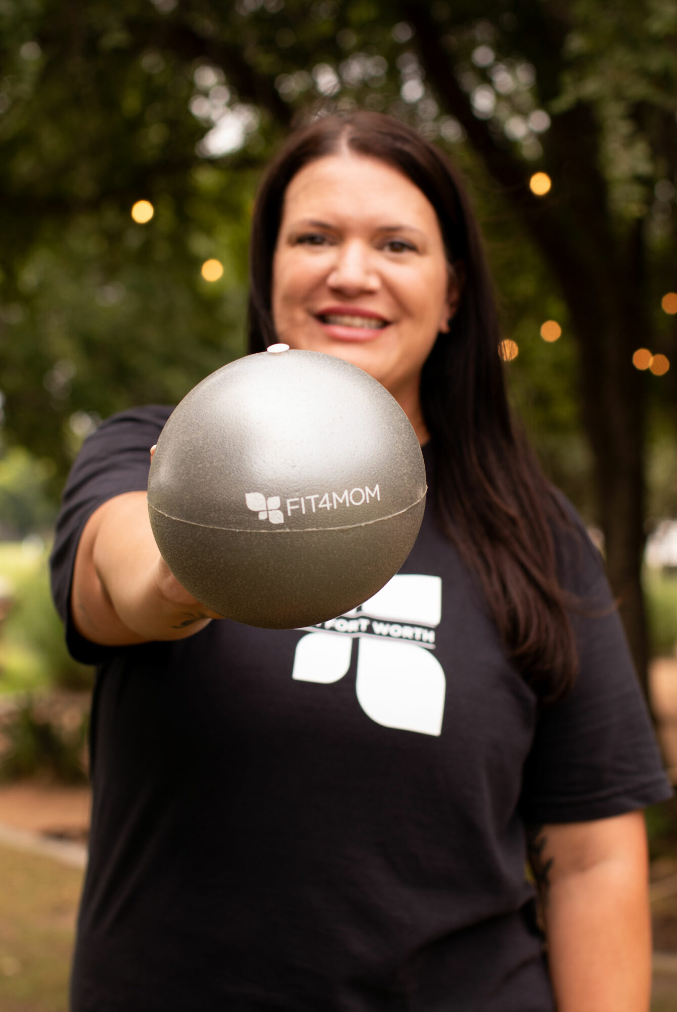 Woman smiling outdoors holding a gray exercise ball with a logo, trees and string lights in background.