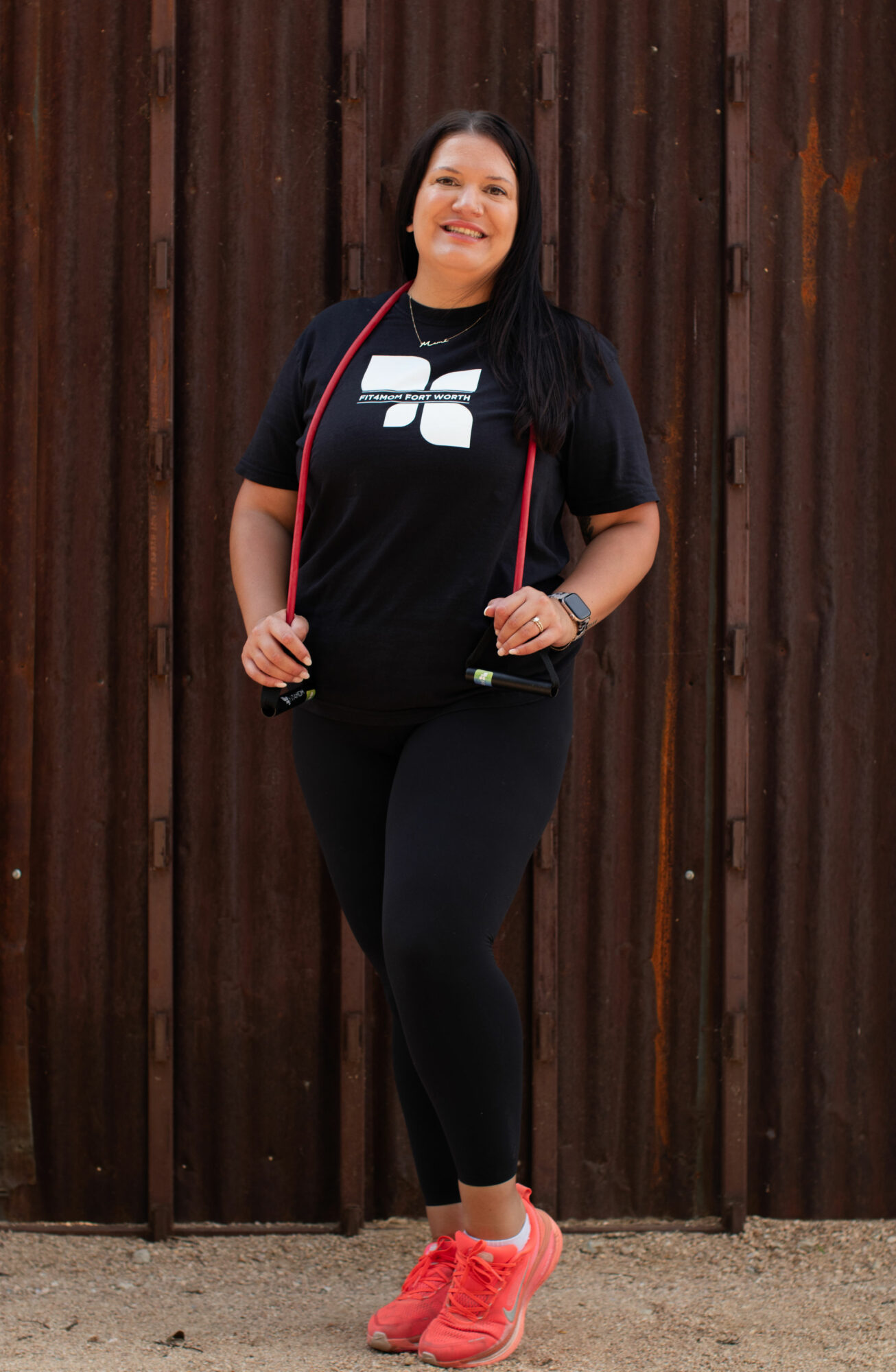 Woman standing outdoors in front of a wooden fence, smiling, wearing a black t-shirt, black leggings, and pink sneakers.