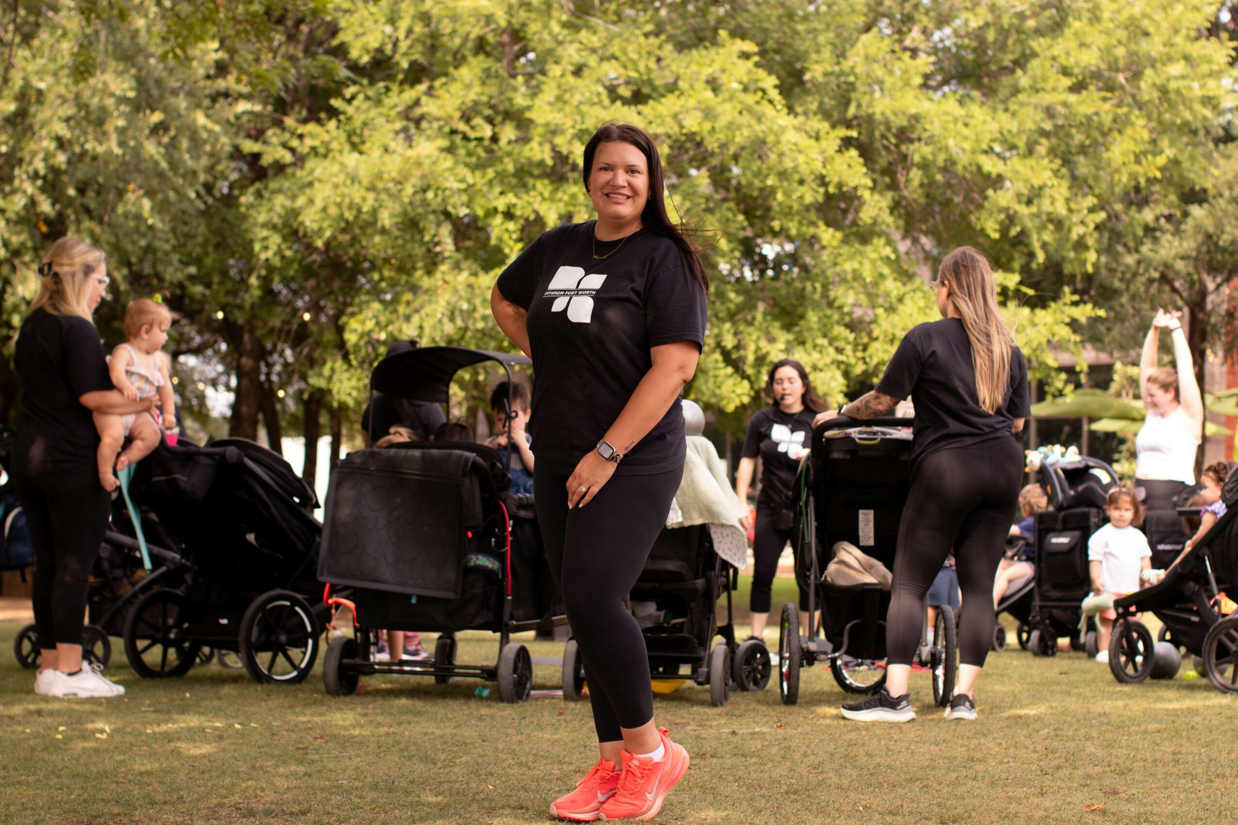 Woman standing outdoors with trees and people in the background, smiling, wearing black clothing and pink shoes.