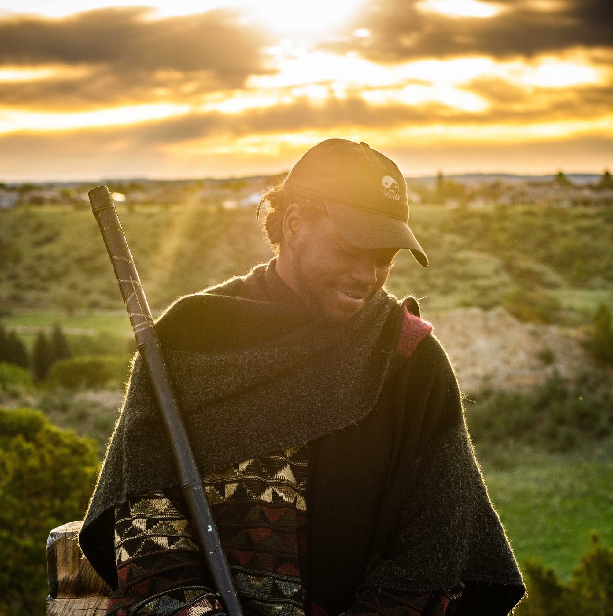 Person wearing a cap and scarf, holding a walking stick, outdoors during sunset with a landscape background.