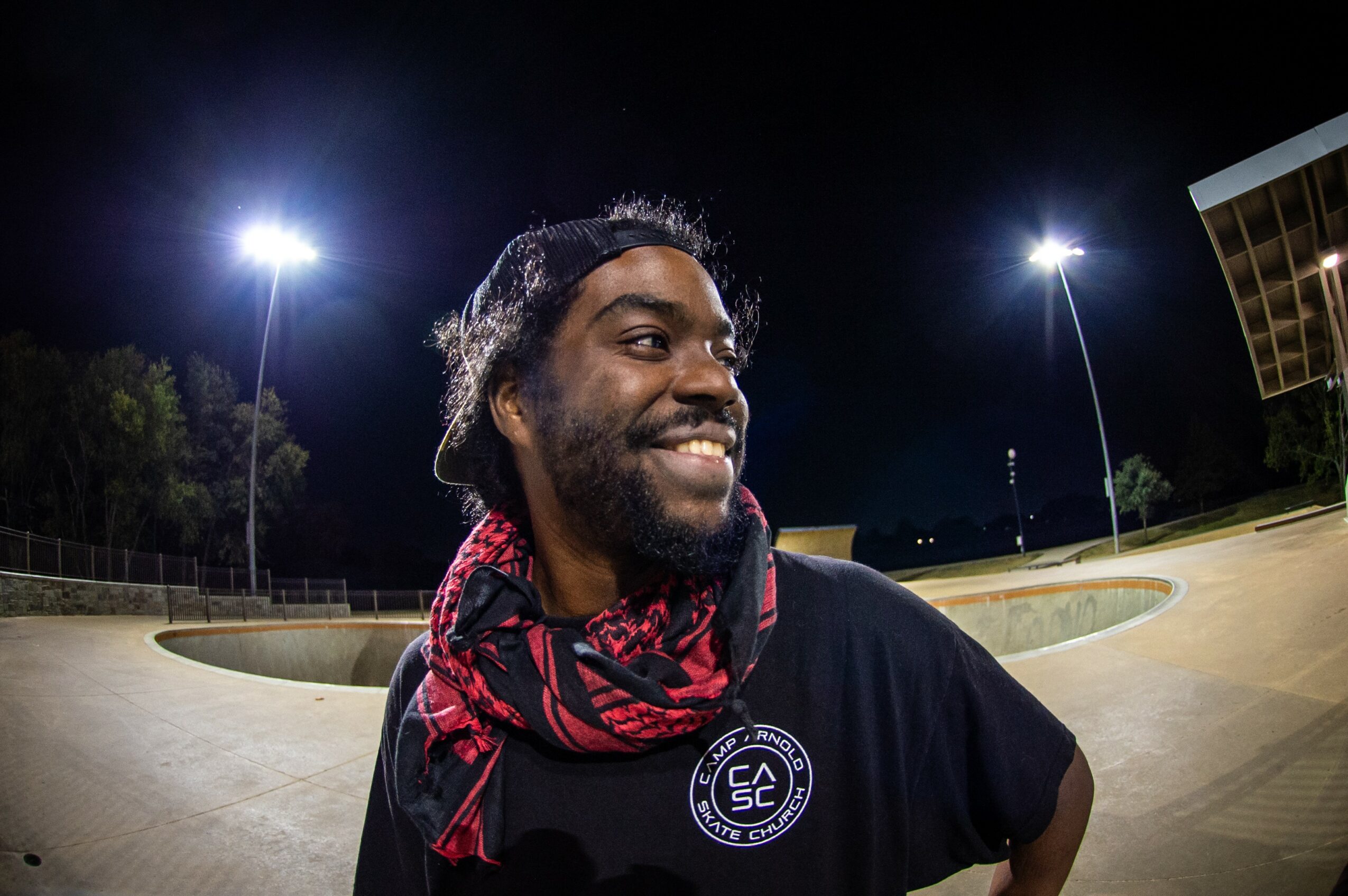 Smiling person with a beard and curly hair, wearing a black shirt and red scarf, outdoors at night with bright lights.