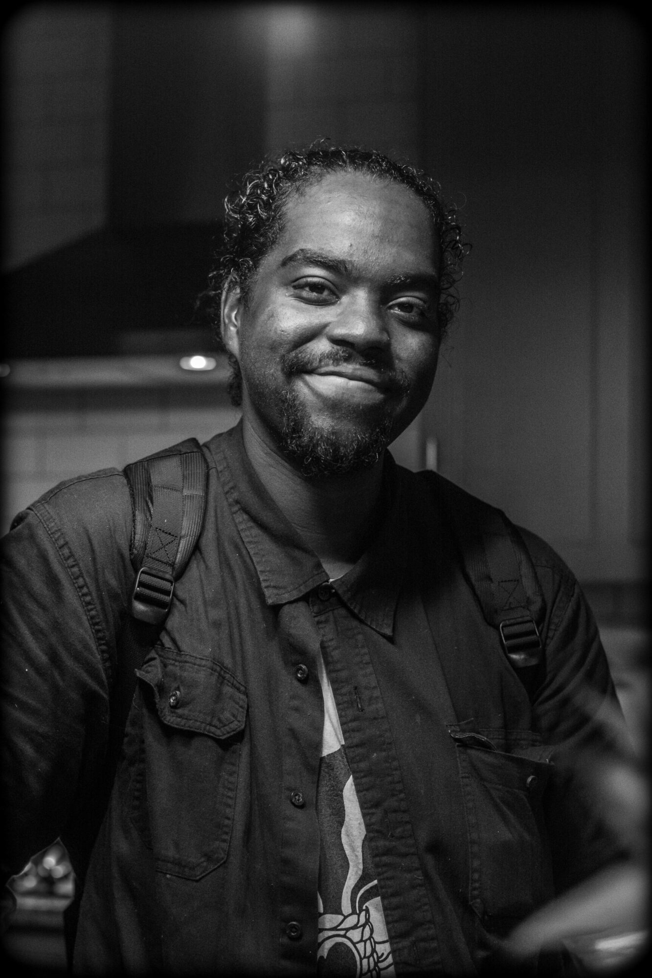 Man with curly hair and beard smiling, wearing a dark jacket and backpack, in an indoor setting.