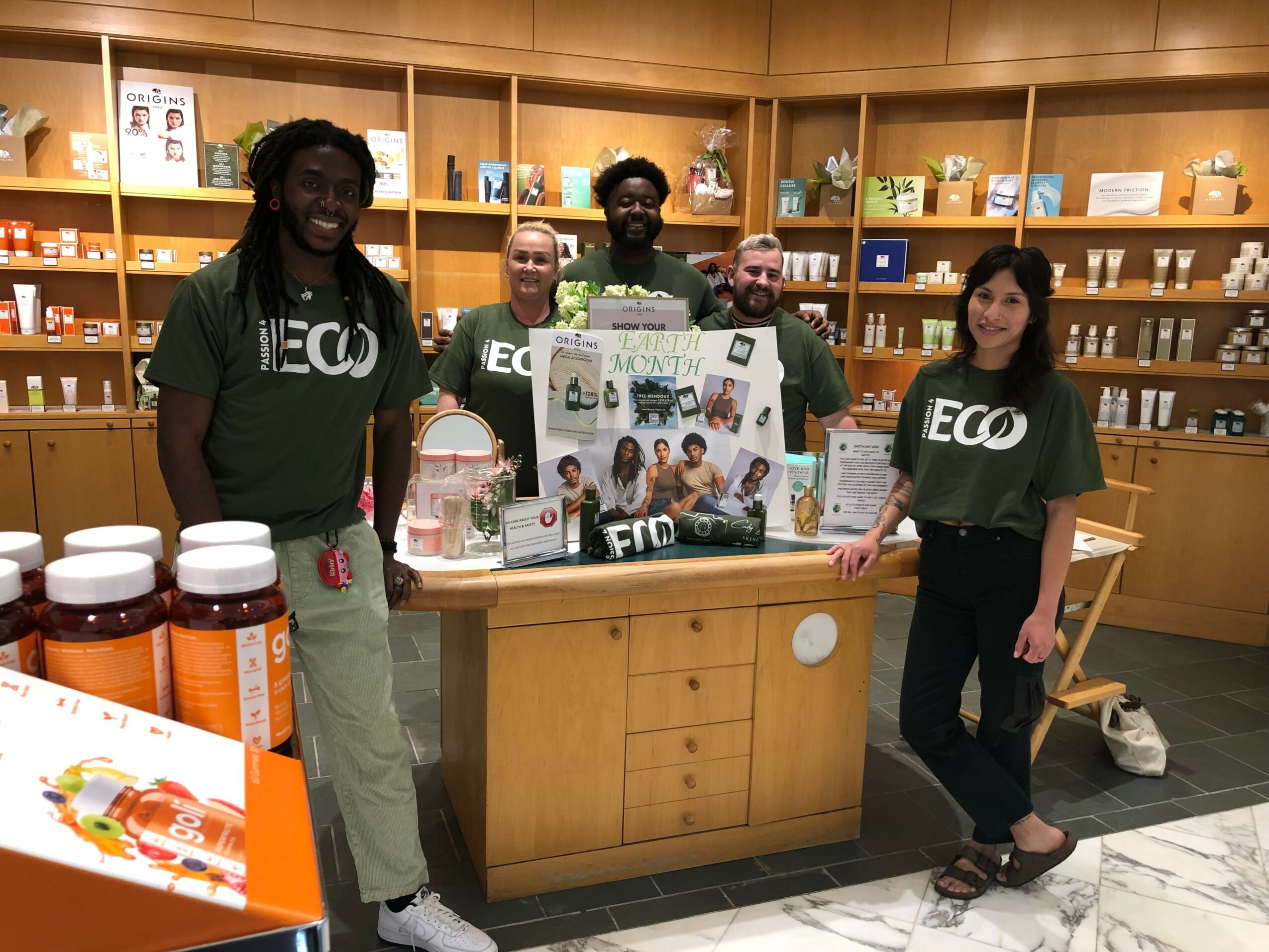Five people stand behind a display table in a store, smiling, with shelves of products in the background.