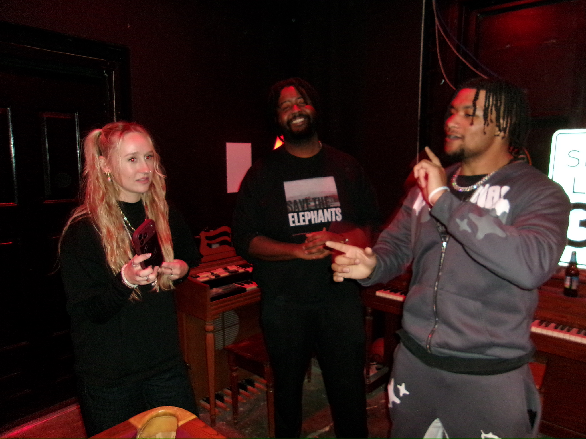 Three people standing indoors, two men and one woman, engaged in conversation, with musical equipment in the background.