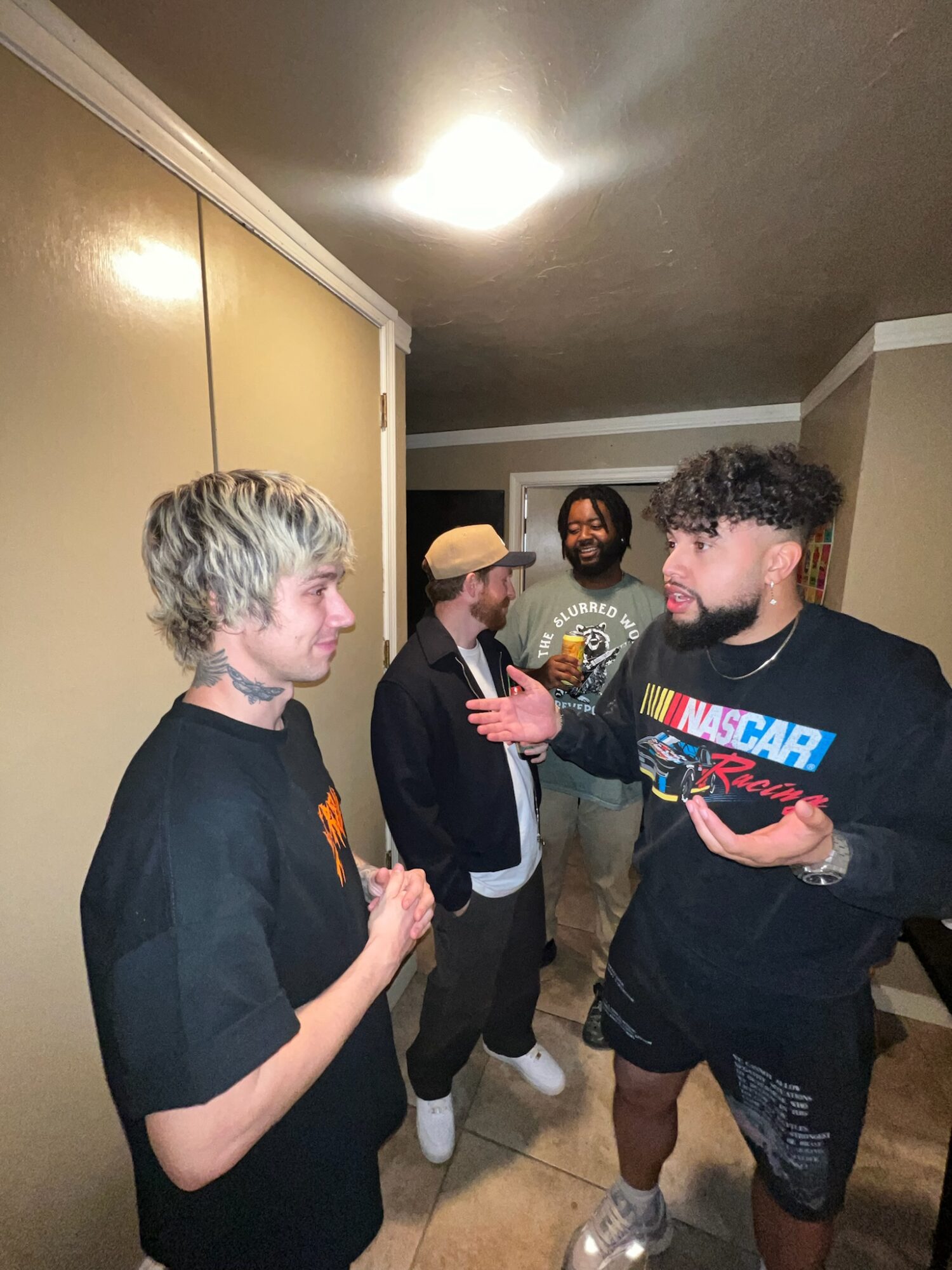 Four people standing indoors, engaged in conversation, with a plain wall and ceiling light visible in the background.