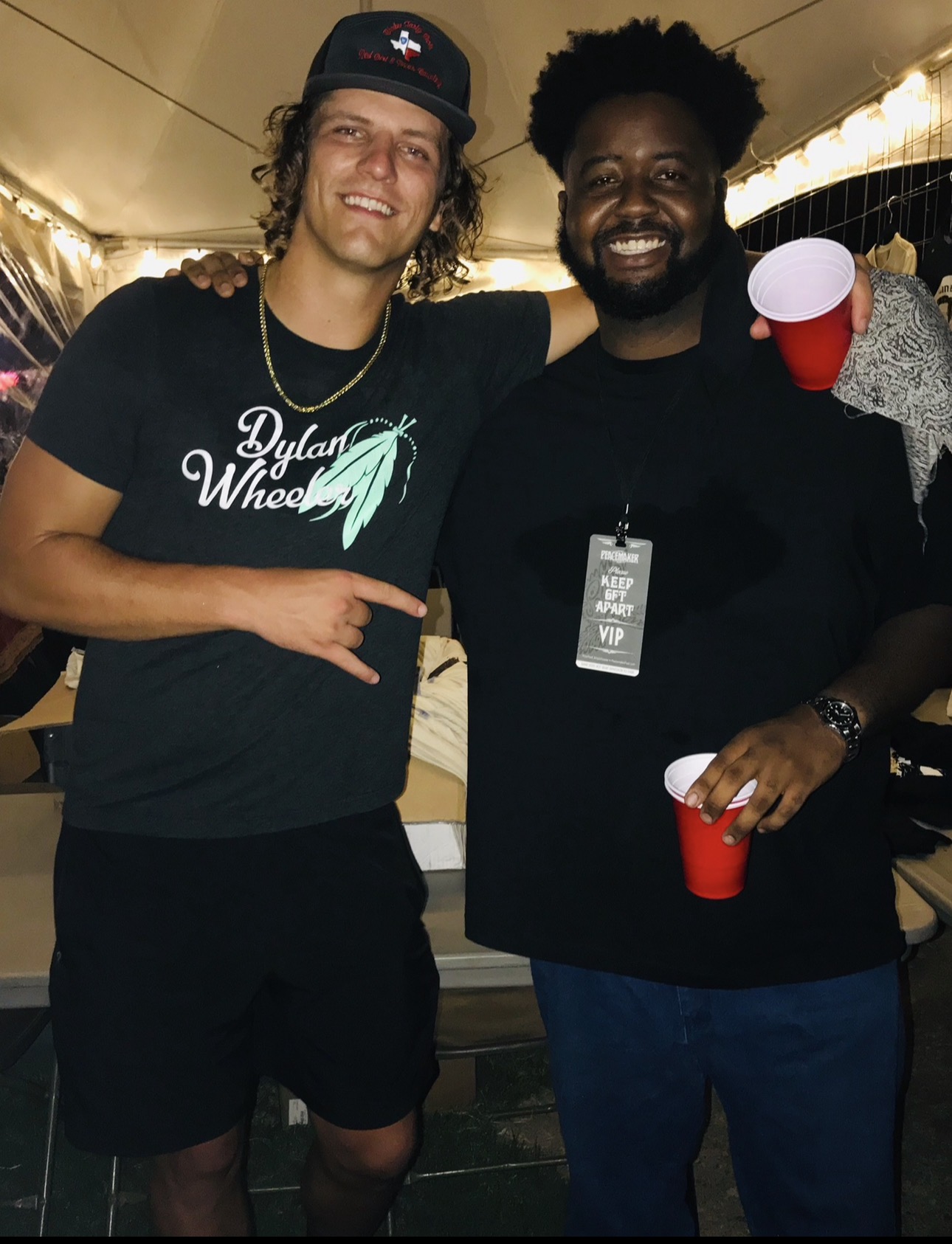 Two men smiling, one holding a red cup, in a party setting with warm lighting.