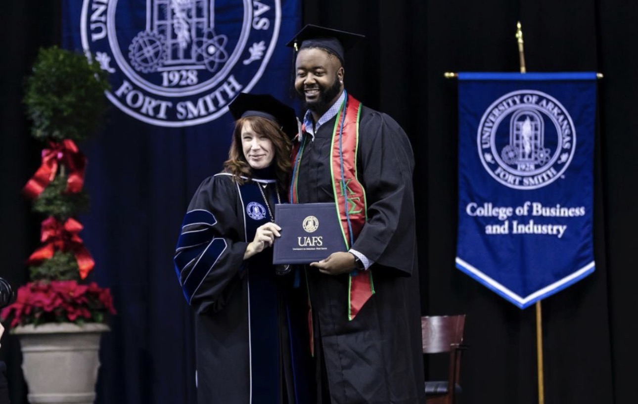 Graduate in cap and gown receiving diploma from a smiling man on stage at graduation ceremony.