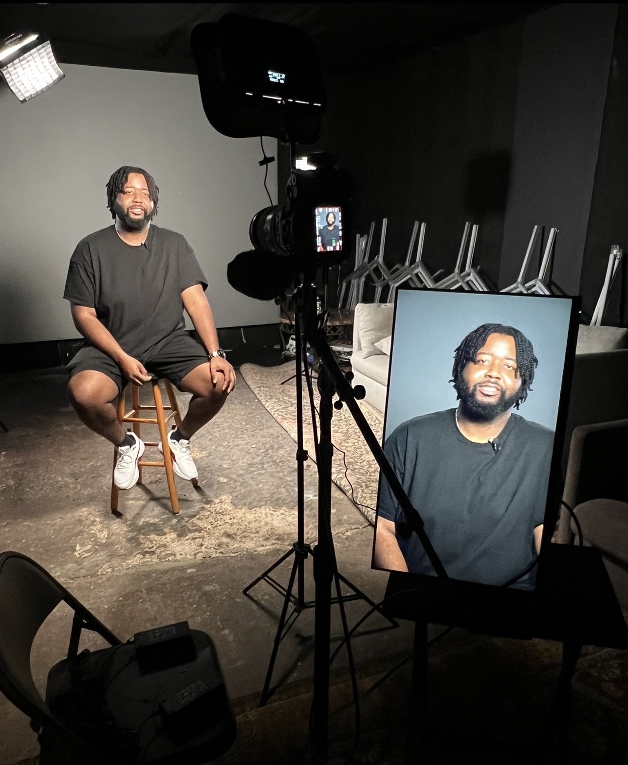 Person sitting on a stool in a studio, being filmed with a camera and lighting, with a monitor showing their image.